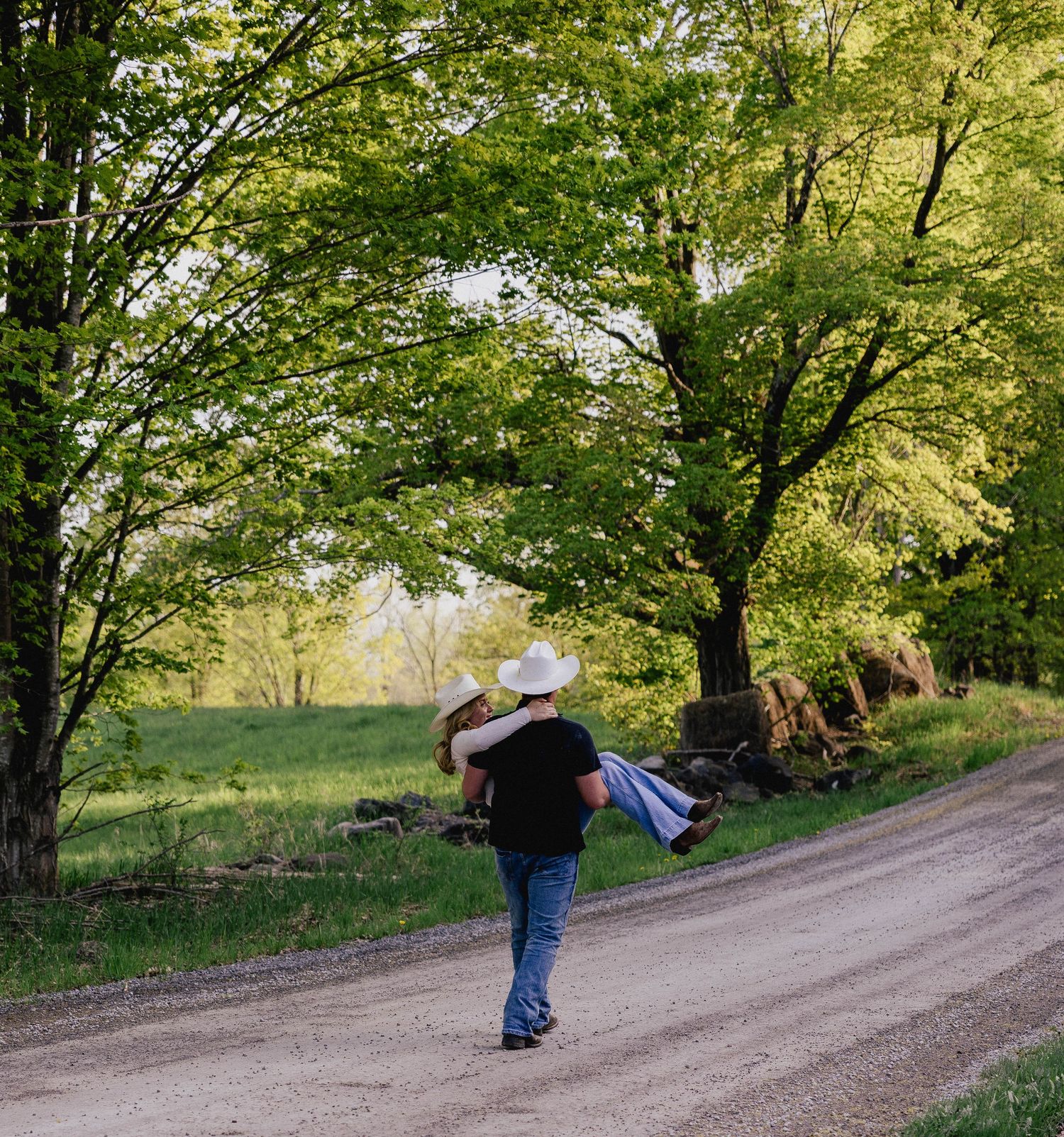 Man carrying woman on dirt road surrounded by lush green trees, romantic country engagement photo.