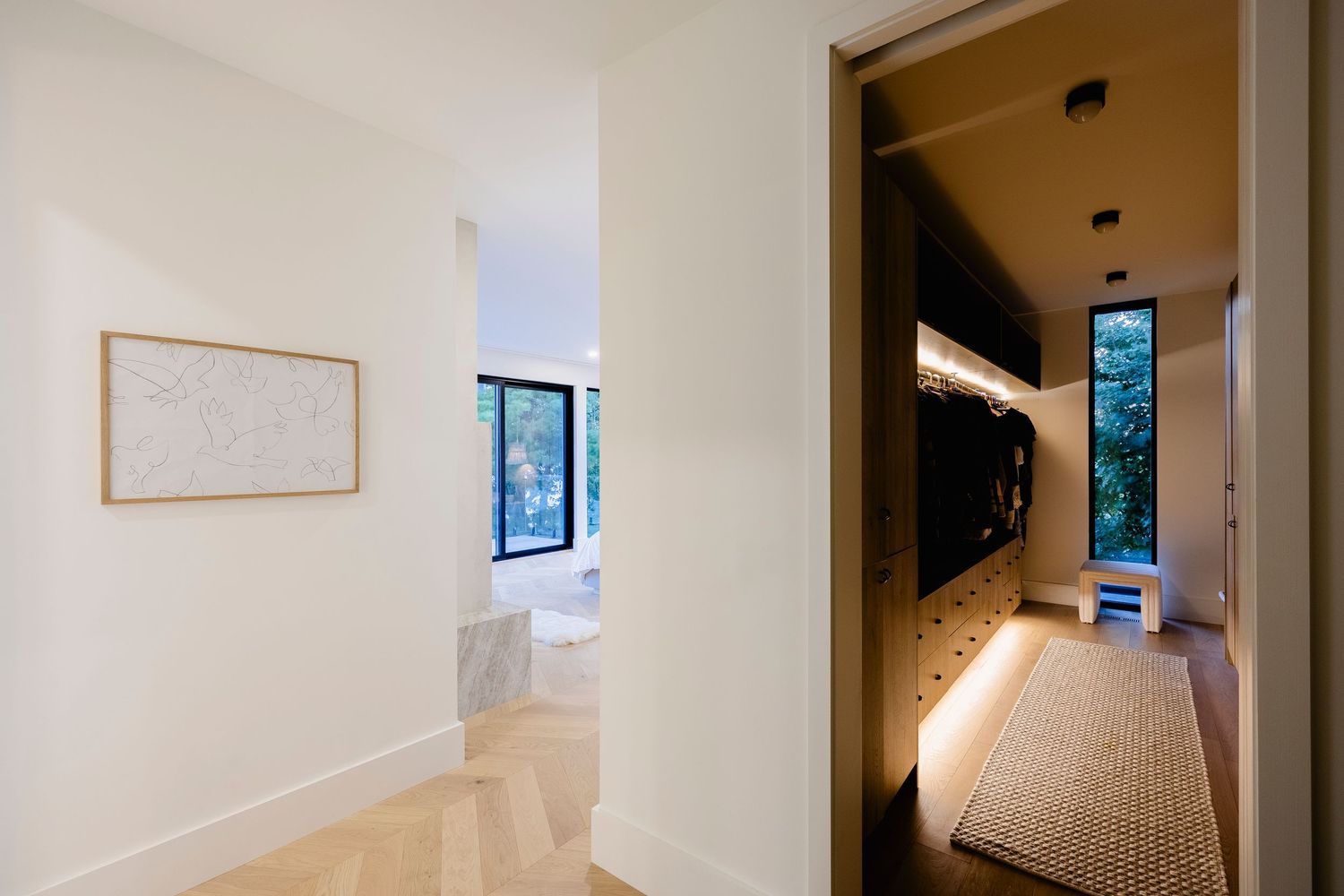 Minimalist hallway with herringbone floors, framed art, and illuminated walk-in mudroom with built-in storage.