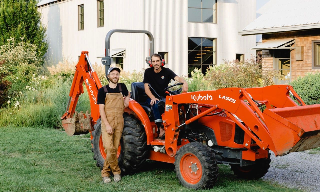 Two people posing beside an orange Kubota tractor in front of a modern white farmhouse.