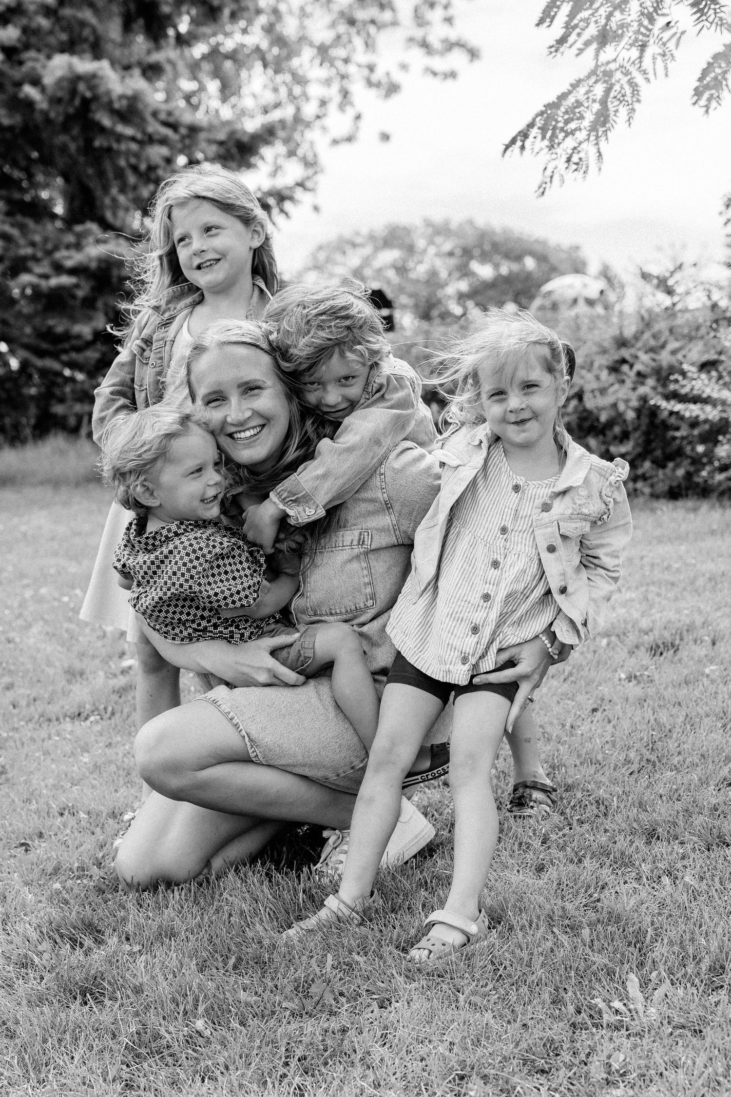 Black and white photo of a smiling mother kneeling on grass, hugging four young blonde children outdoors.
