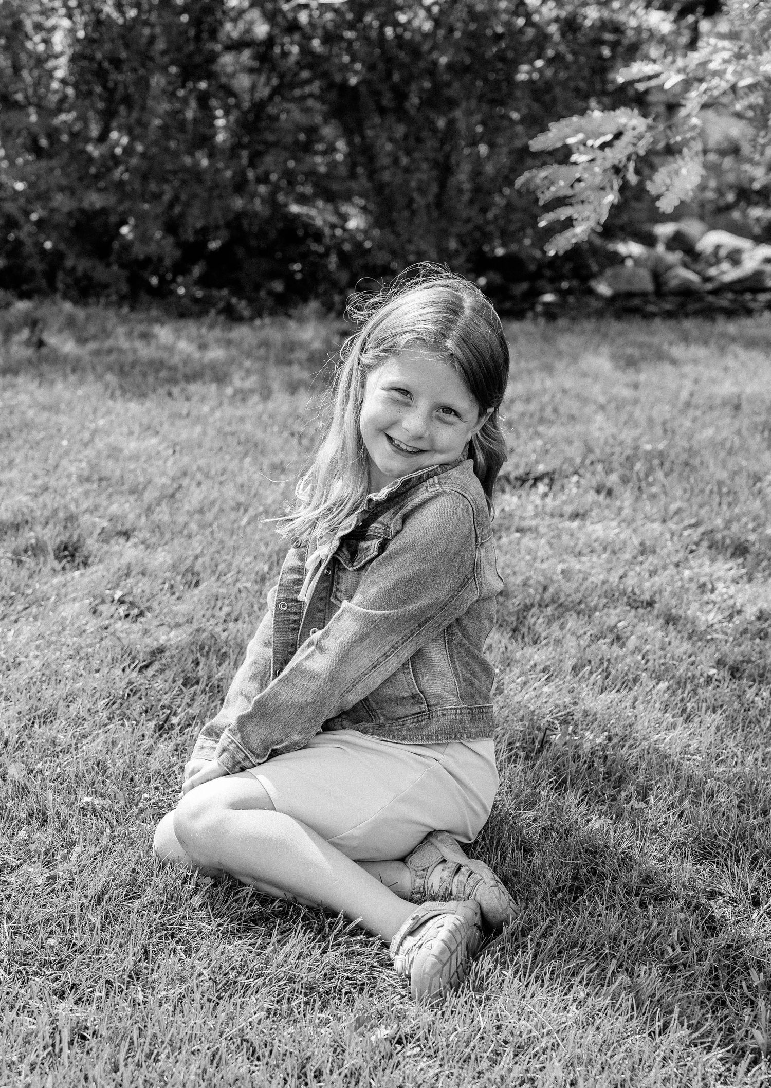 A smiling young girl with long hair sits cross-legged on green grass in a garden, wearing a denim jacket, in black and white.