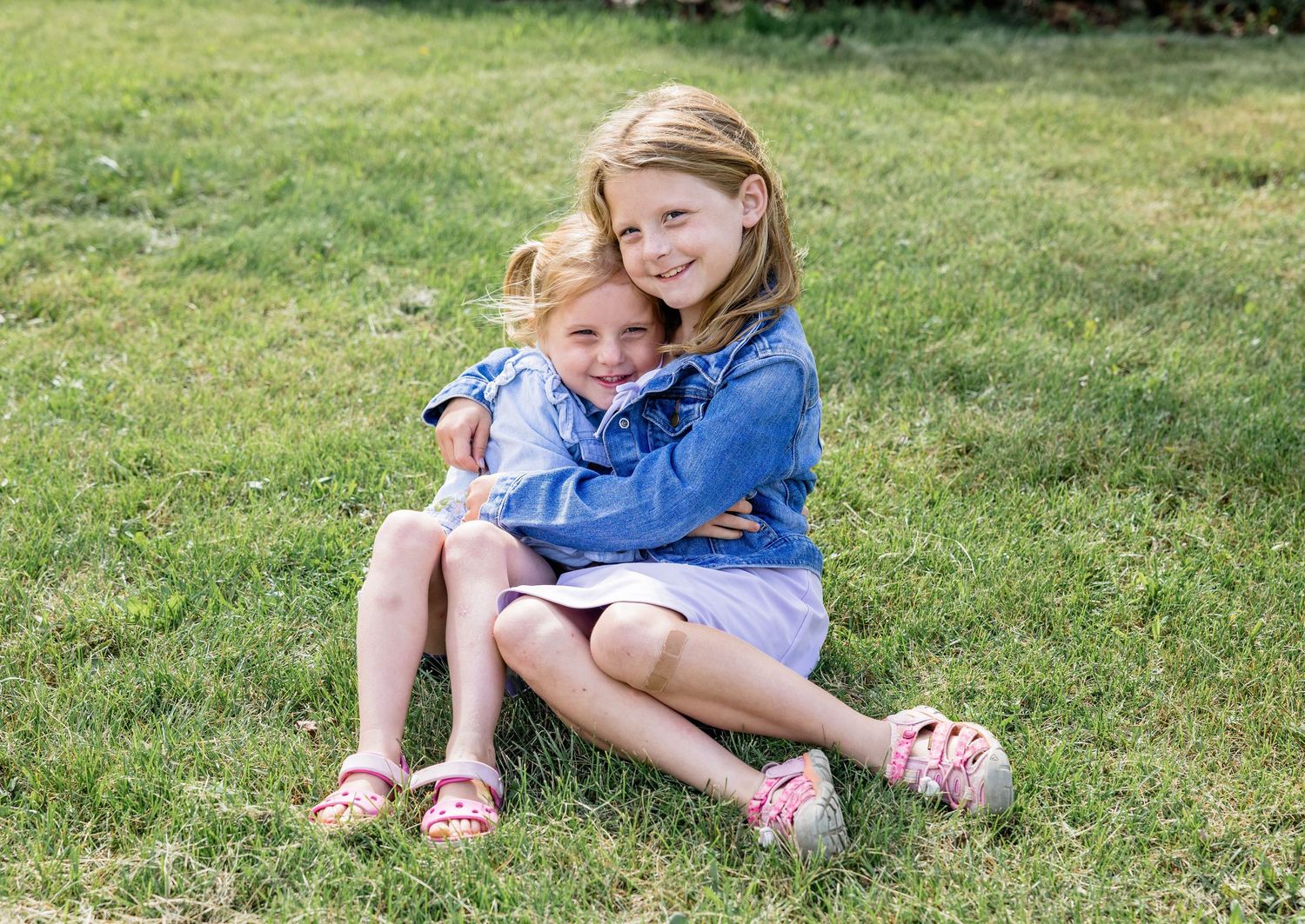 Two young girls sitting on green grass, hugging and smiling, wearing matching blue outfits and pink sandals.