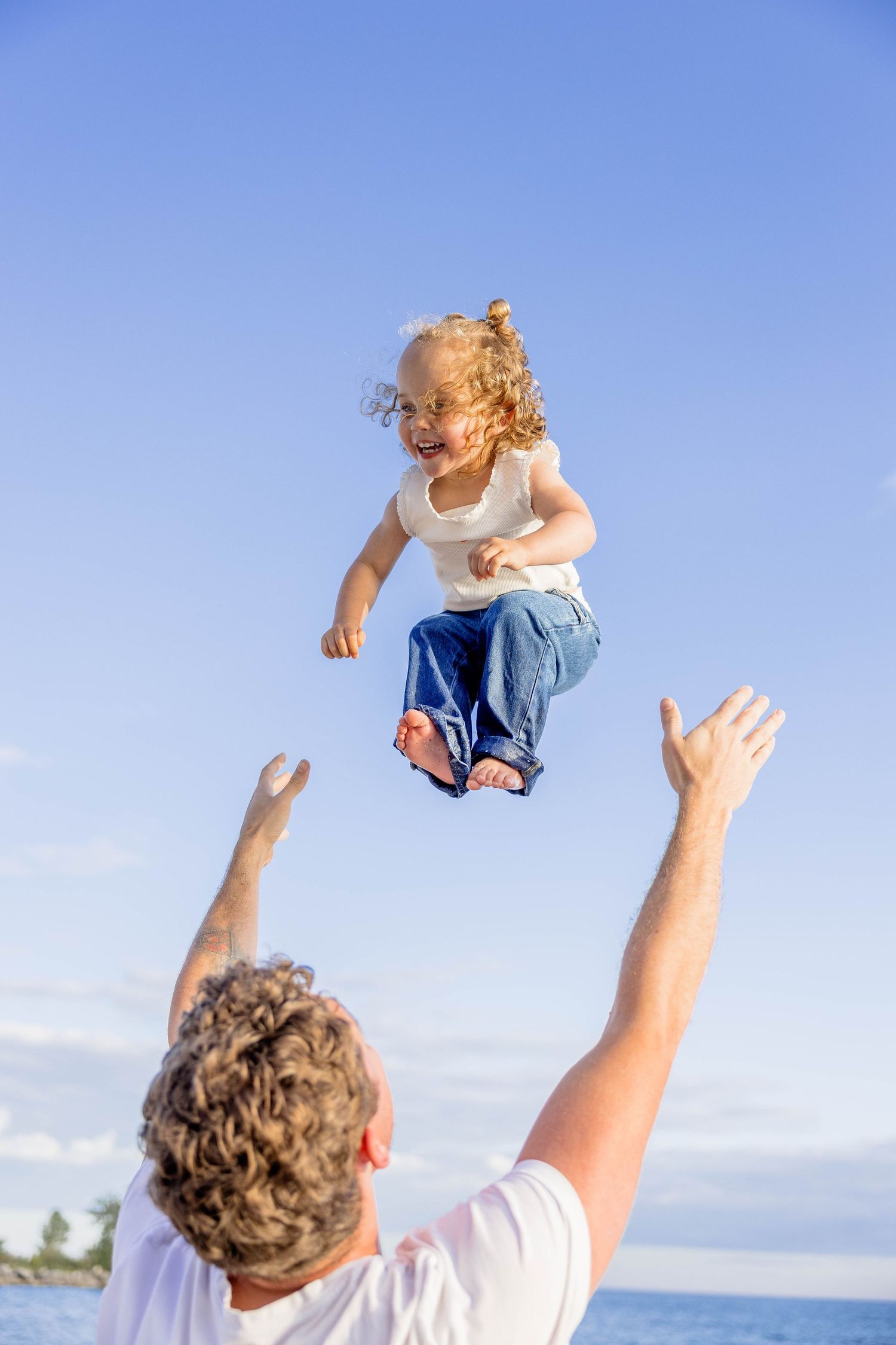 Father tossing laughing curly-haired toddler in the air at the beach against a clear blue sky.