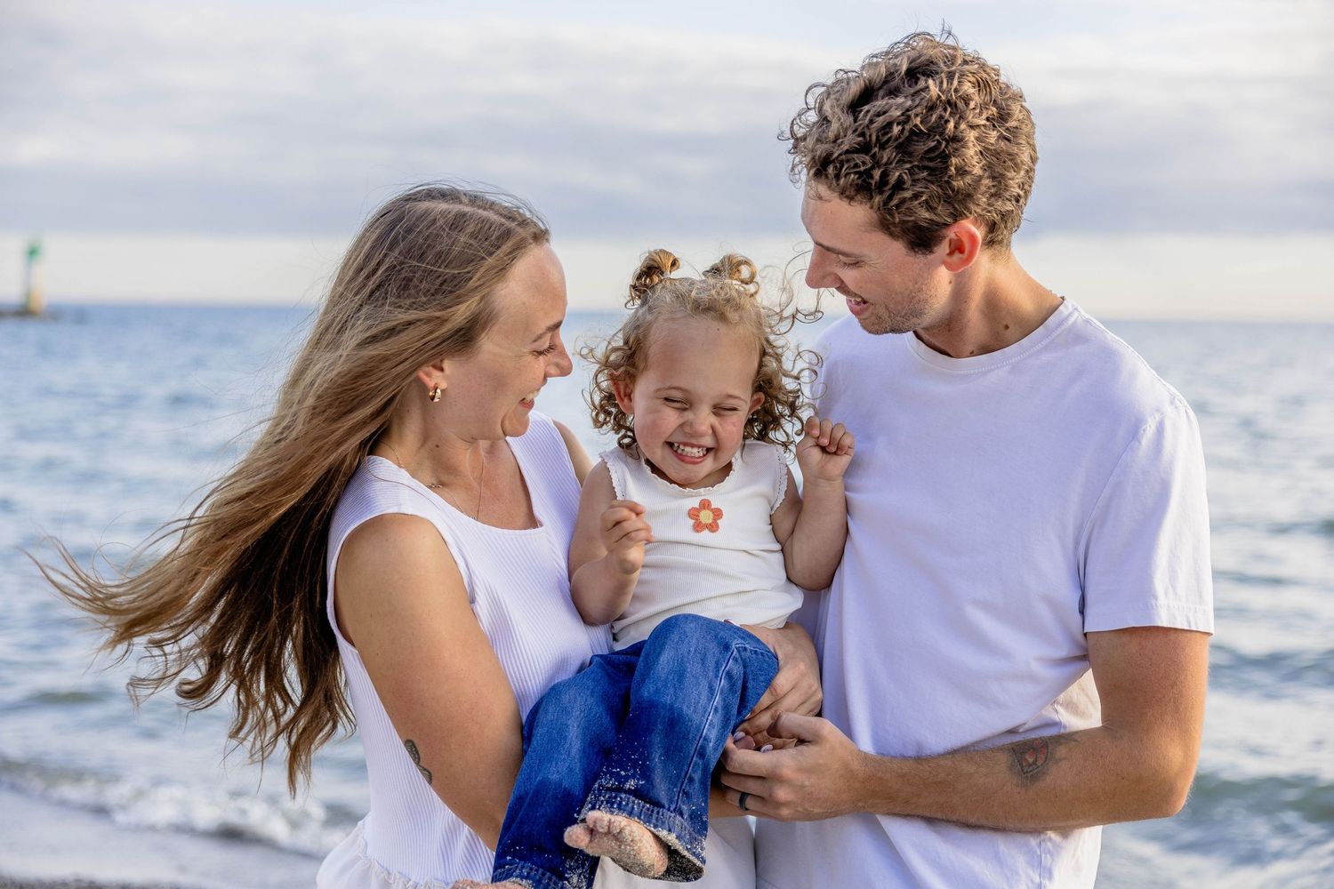 Happy family with parents holding smiling toddler girl at beach with ocean waves in background.