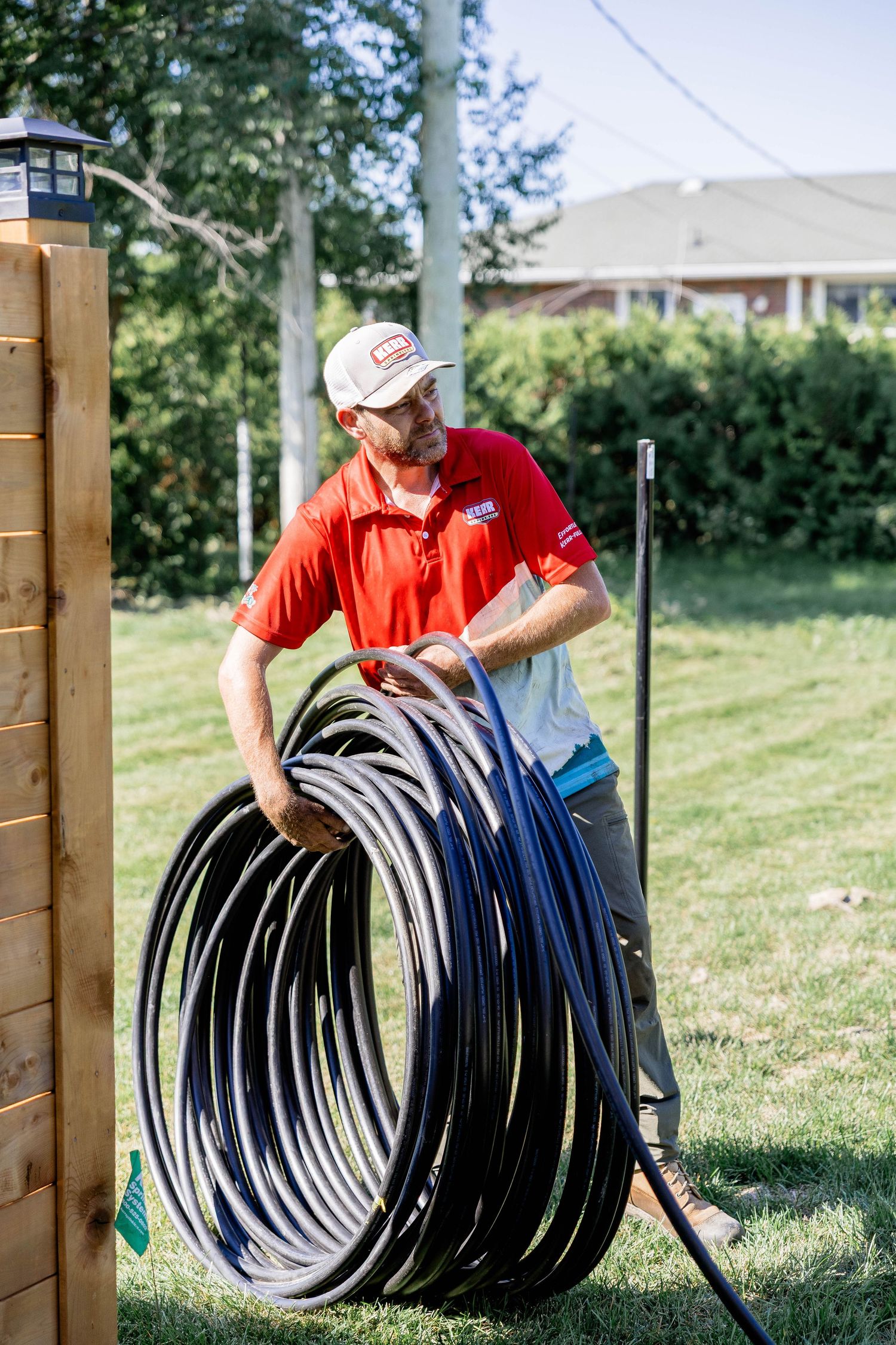 Technician in red polo carries large coiled black irrigation pipe in a residential backyard on a sunny day.
