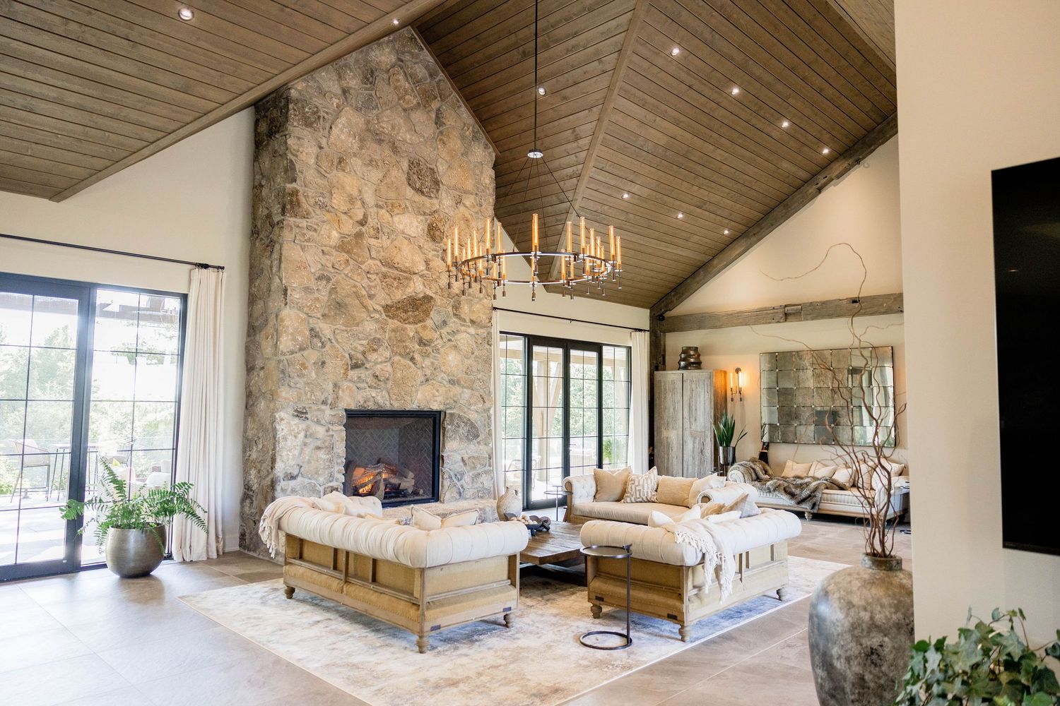 Luxury rustic living room with soaring stone fireplace, wood ceiling, chandeliers, and large windows.