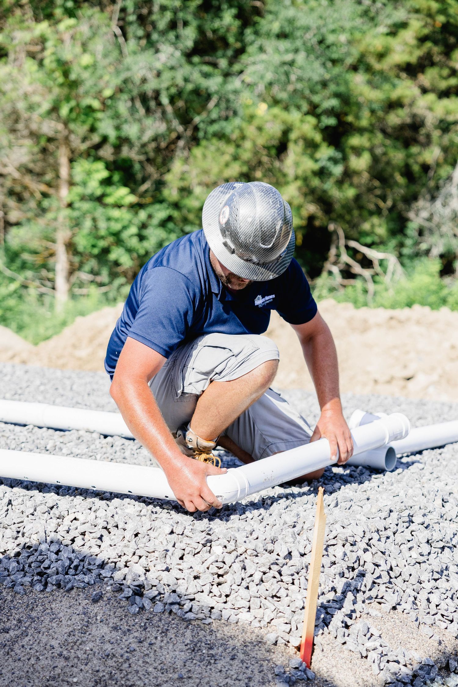 Construction worker in hard hat installs PVC pipes on gravel foundation at building site.