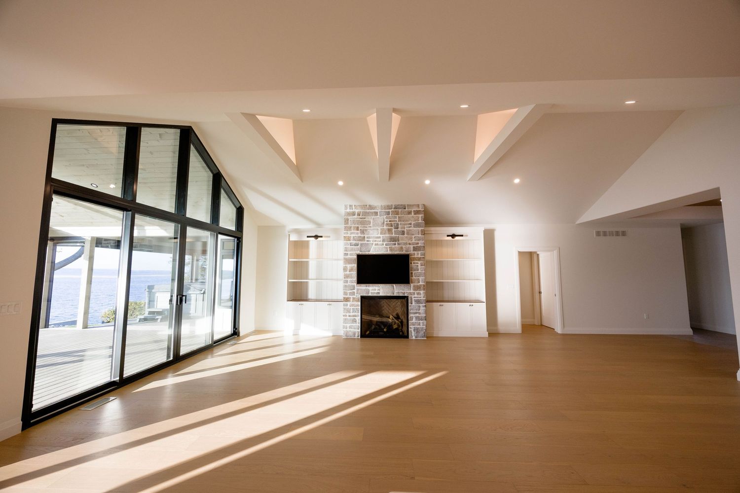 Spacious empty living room with vaulted ceilings, large black-framed windows, stone fireplace, and light hardwood floors.