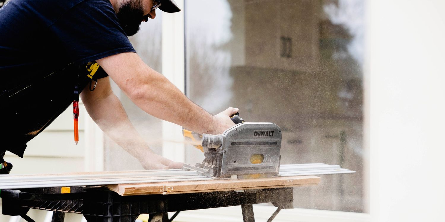 A carpenter uses a circular saw to cut material on a workbench near a window, with sawdust flying.