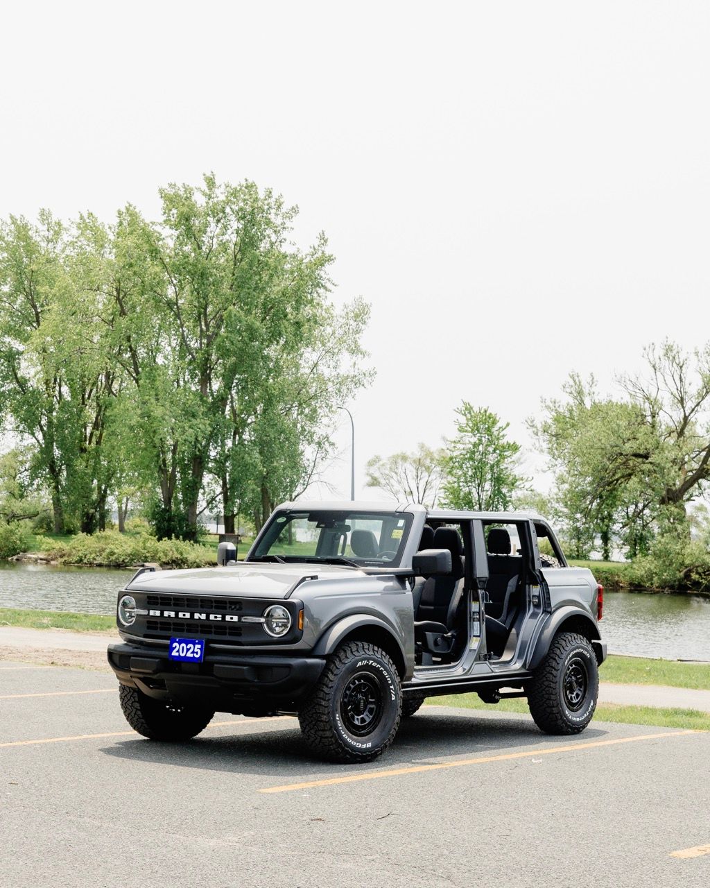 2025 Ford Bronco 4-door in gray with doors off, parked beside a calm waterfront with lush trees.