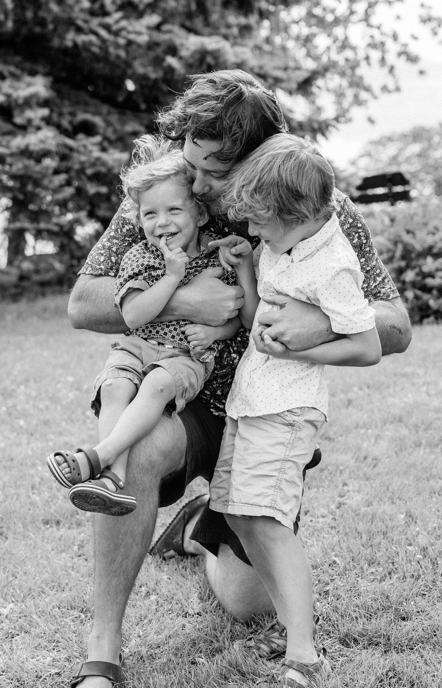 Black and white photo of a father hugging and kissing two young children outdoors on grass with trees in background.