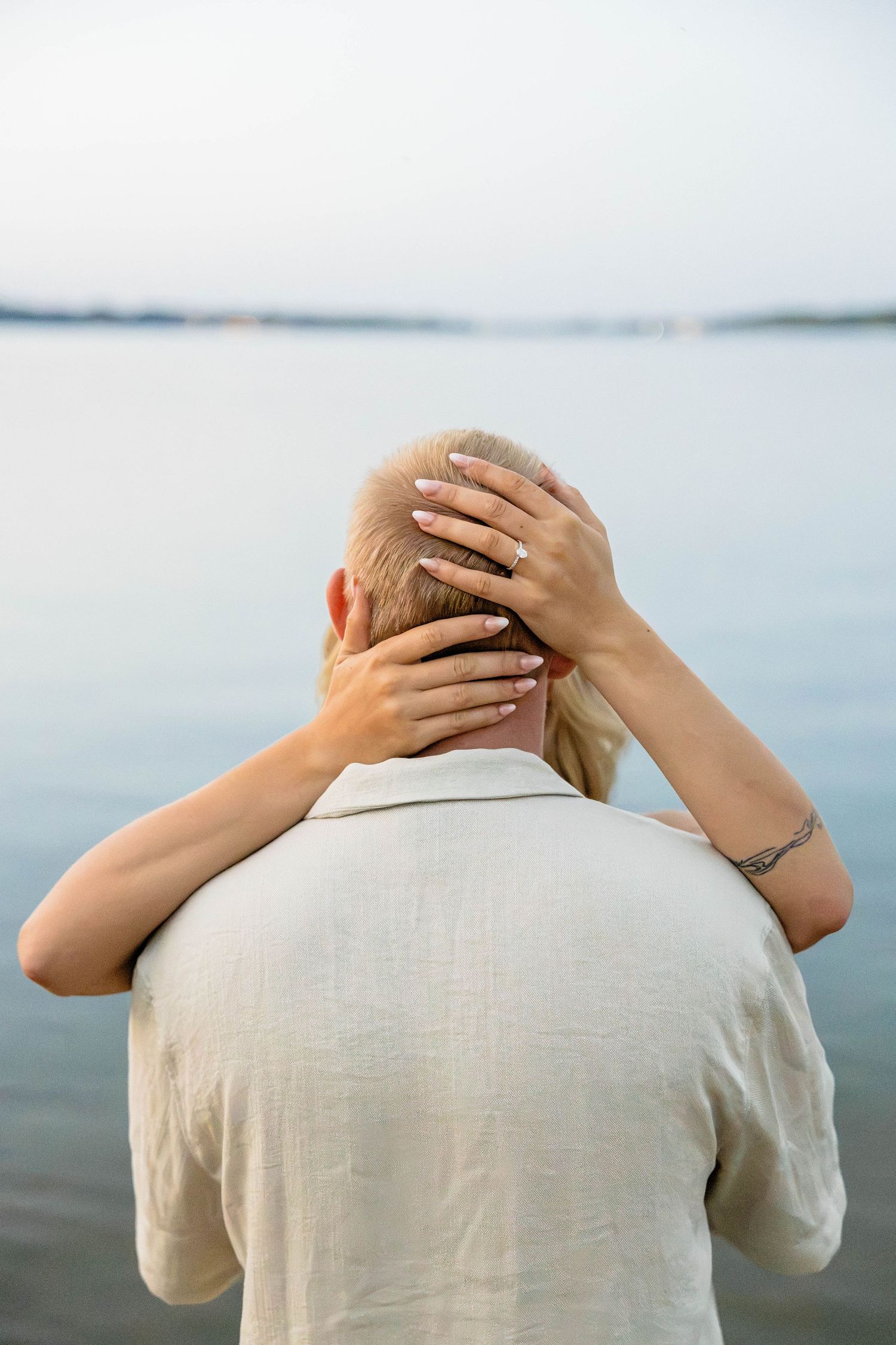 Person with short blonde hair viewed from behind, hands resting on head, gazing at calm water horizon.