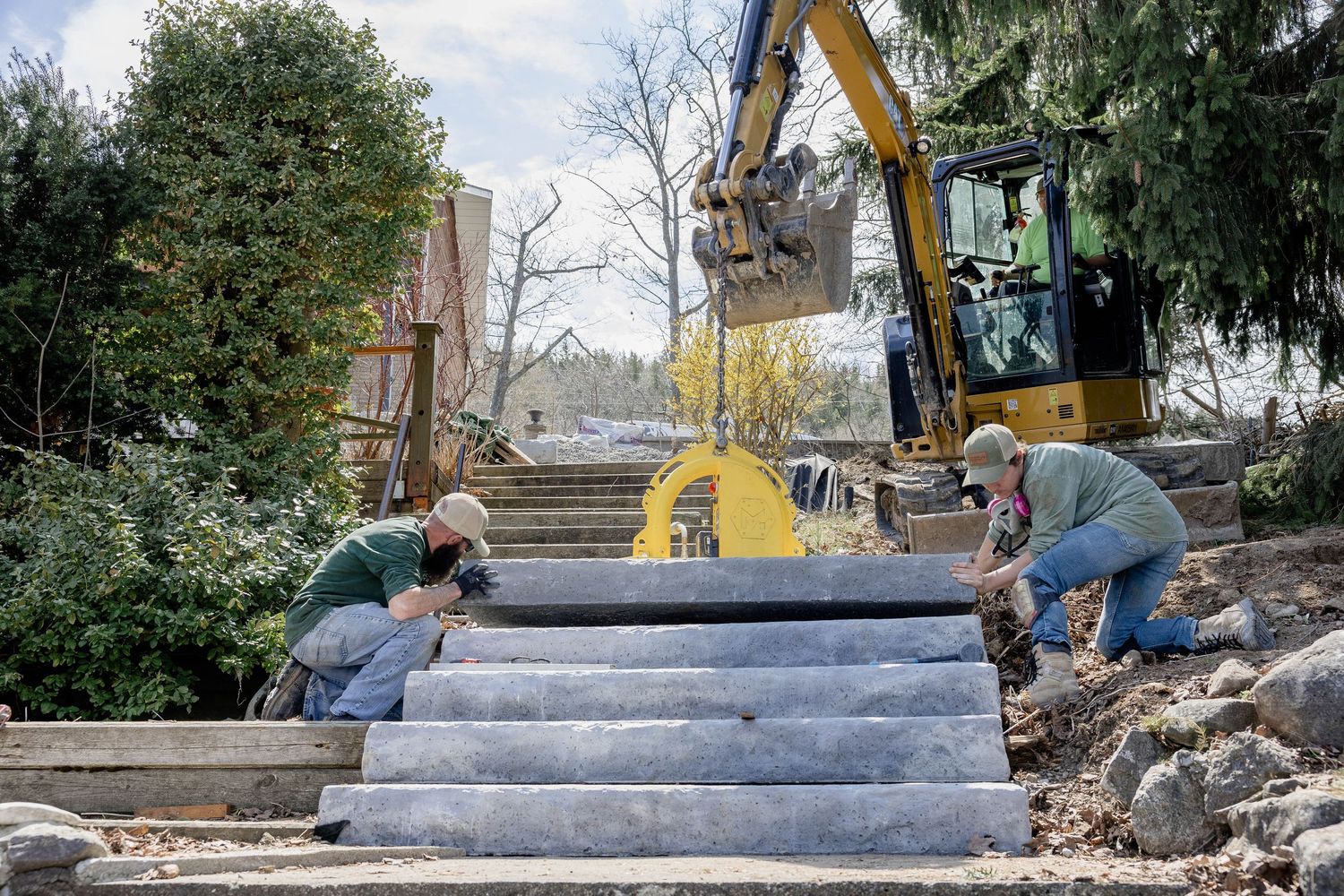 Two workers and an excavator carefully position large concrete steps at an outdoor landscaping site.