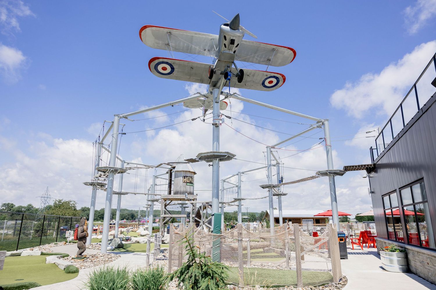 Vintage biplane mounted above an outdoor adventure ropes course attraction on a sunny day.