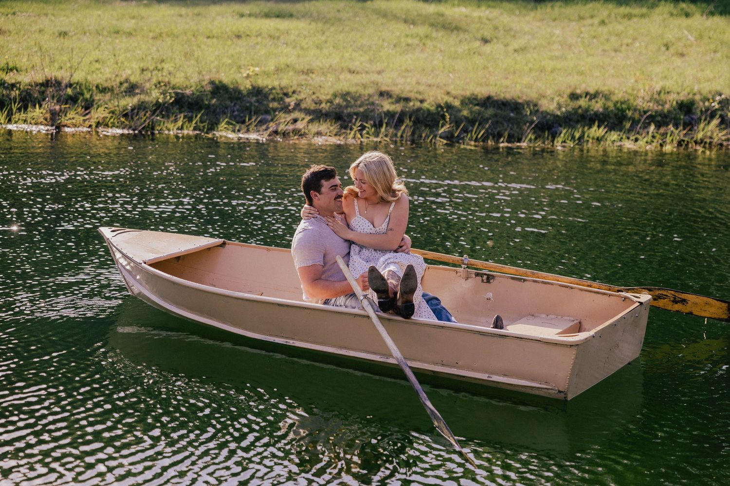 Smiling couple cuddling in a wooden rowboat on a sparkling green lake during golden hour.