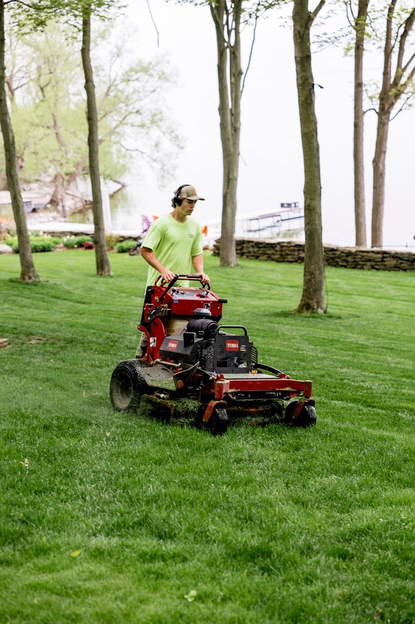 Landscaper operating red Toro stand-on mower on lush green lawn near lake and stone wall.