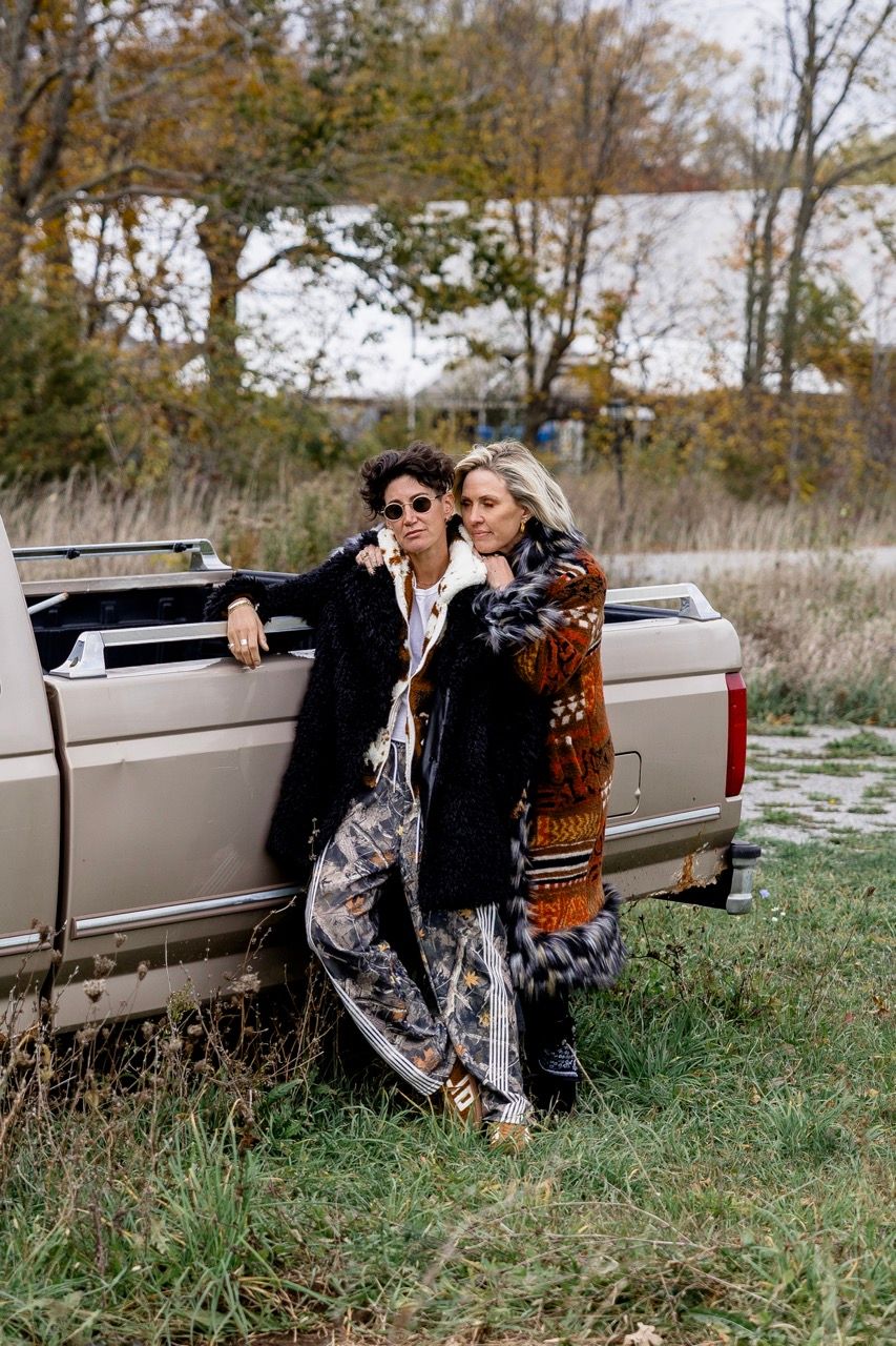Two women in eclectic fall outfits leaning against a vintage pickup truck in autumn landscape.