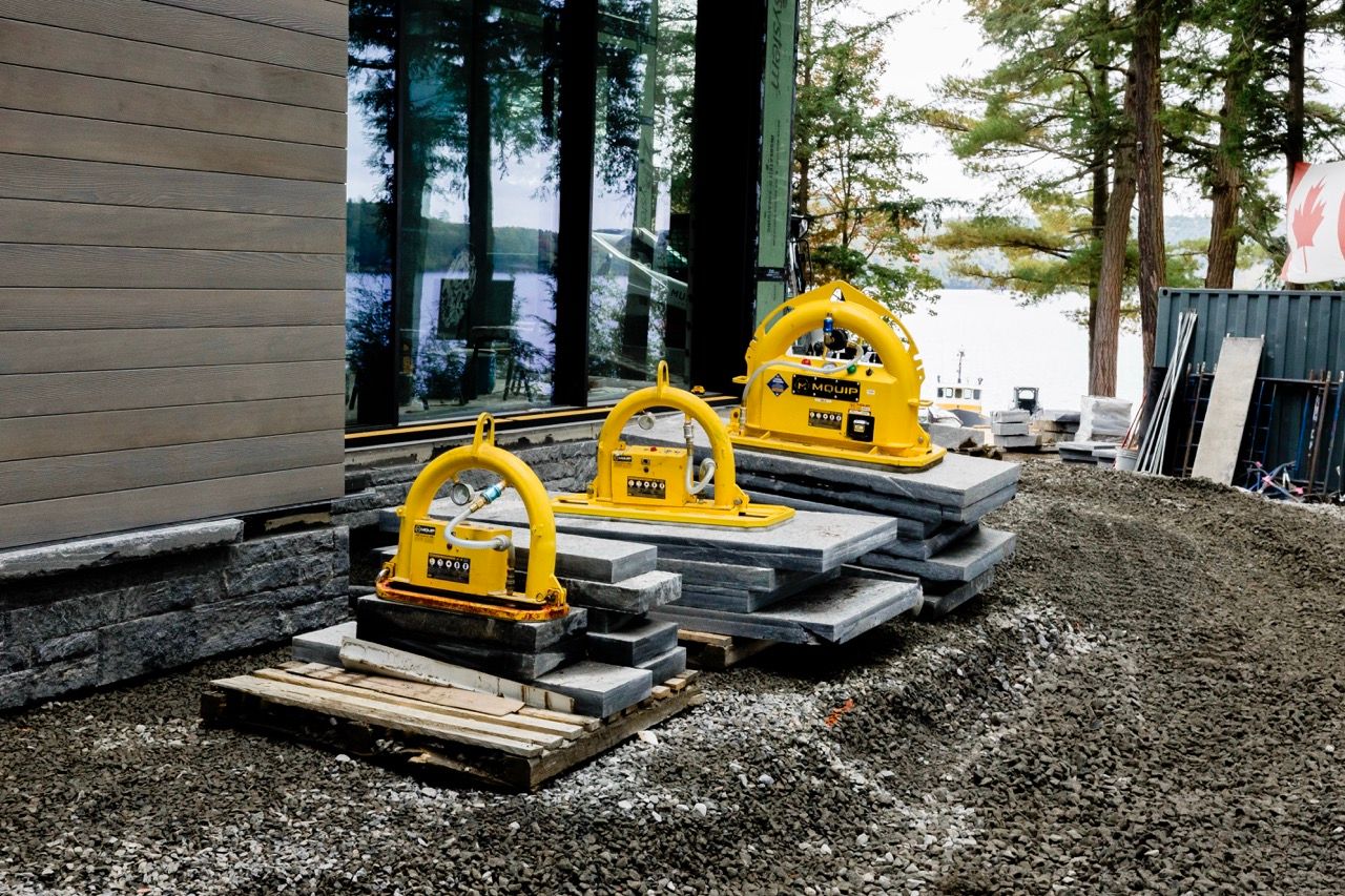 Three yellow MQUIP vacuum stone lifters placed on gravel near a modern lakeside building under construction.