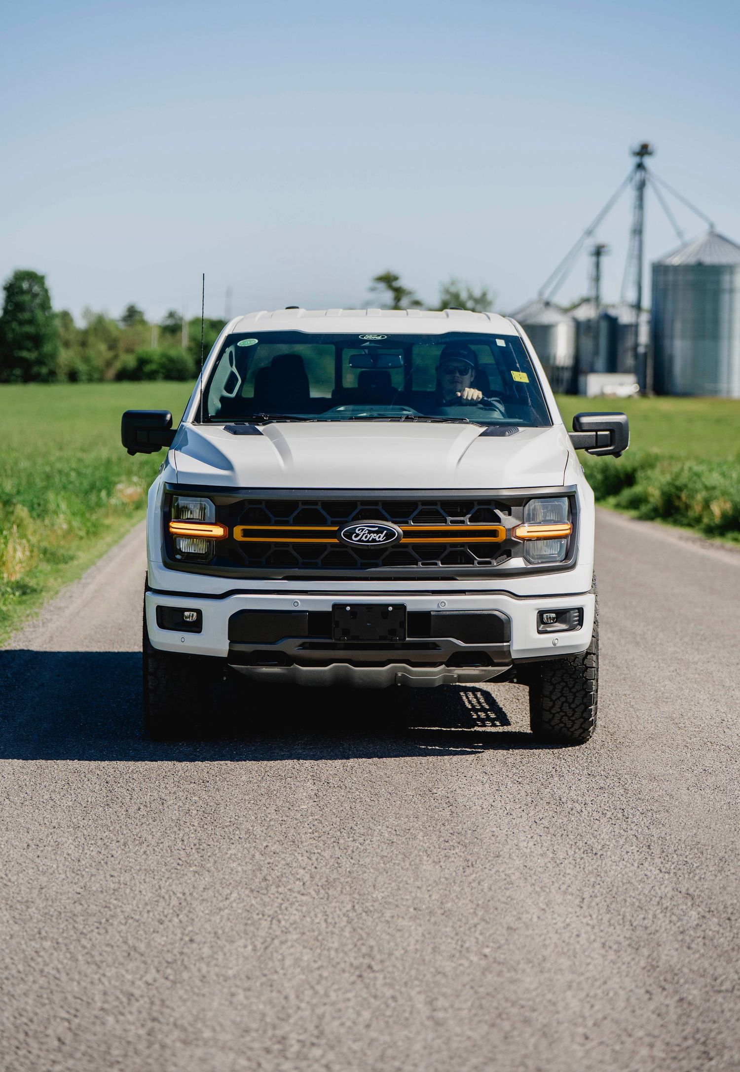 White Ford F-150 truck driving down a rural road with grain silos and green fields in the background.