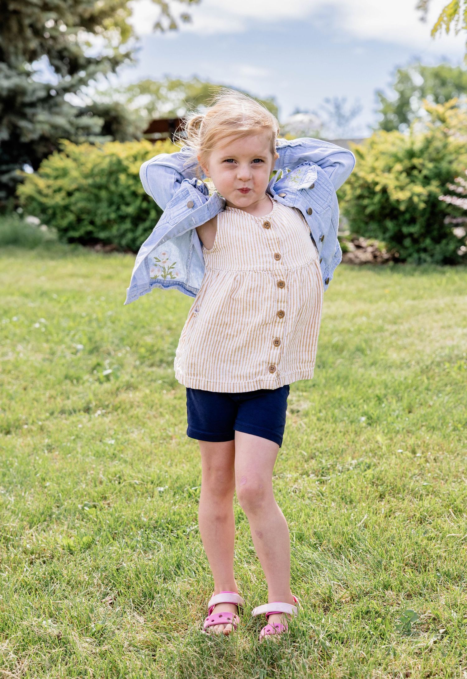 Toddler girl in striped top, navy shorts, and denim jacket poses playfully on green grass in a sunny backyard.