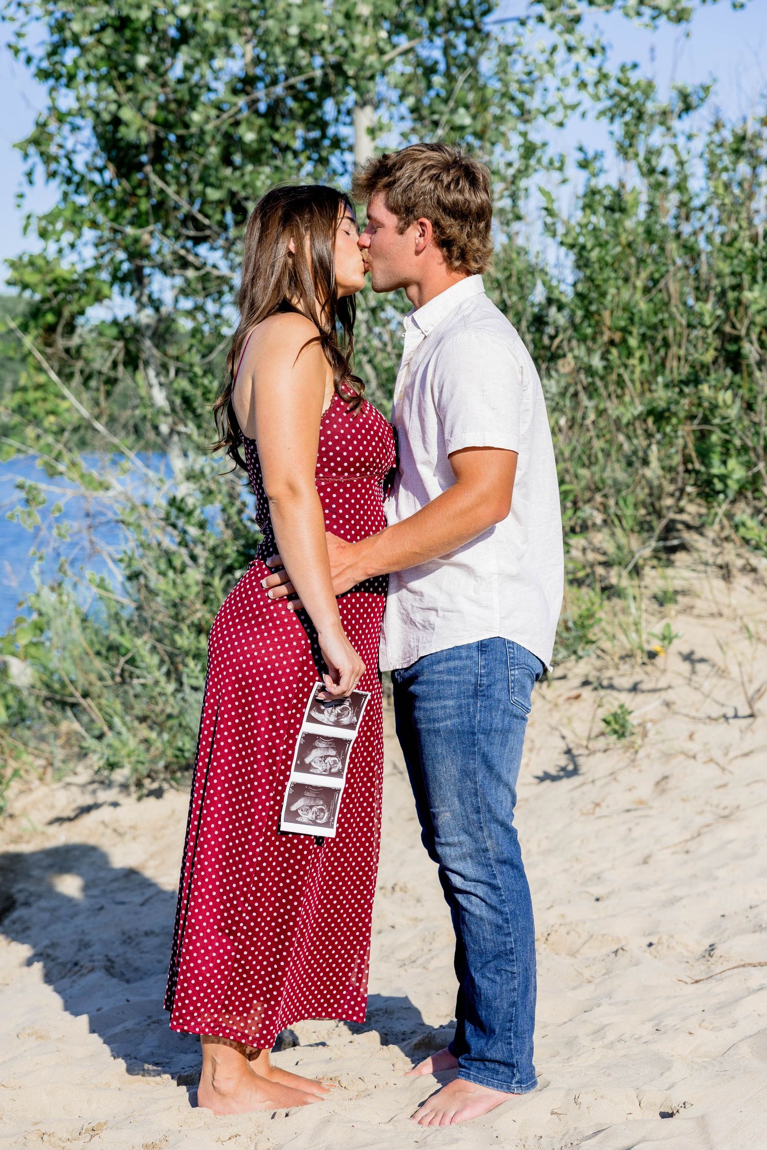 Pregnant couple kissing on sandy beach, woman in red polka dot dress holding ultrasound photos outdoors.