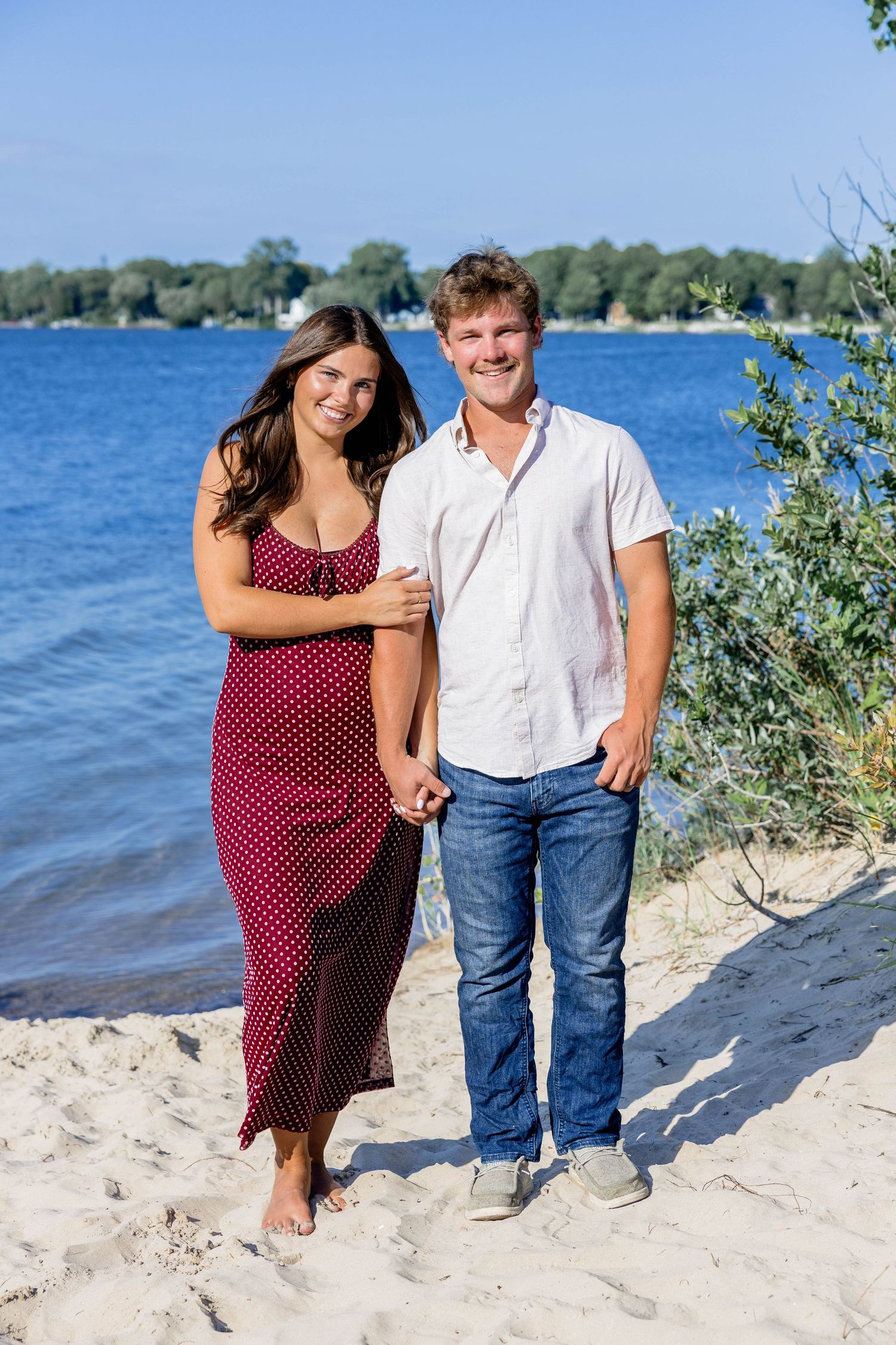 Couple smiling on sandy beach by blue lake, woman in red polka dot dress, man in white shirt and jeans.