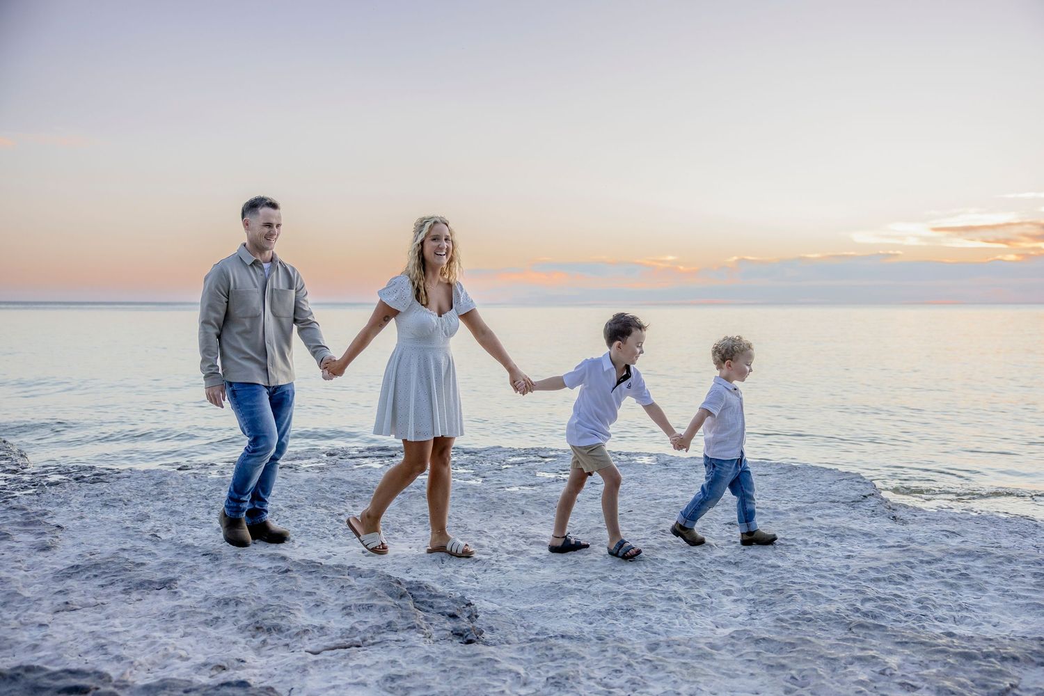 Family of four holding hands walking on rocky beach at sunset with calm ocean waters in background.