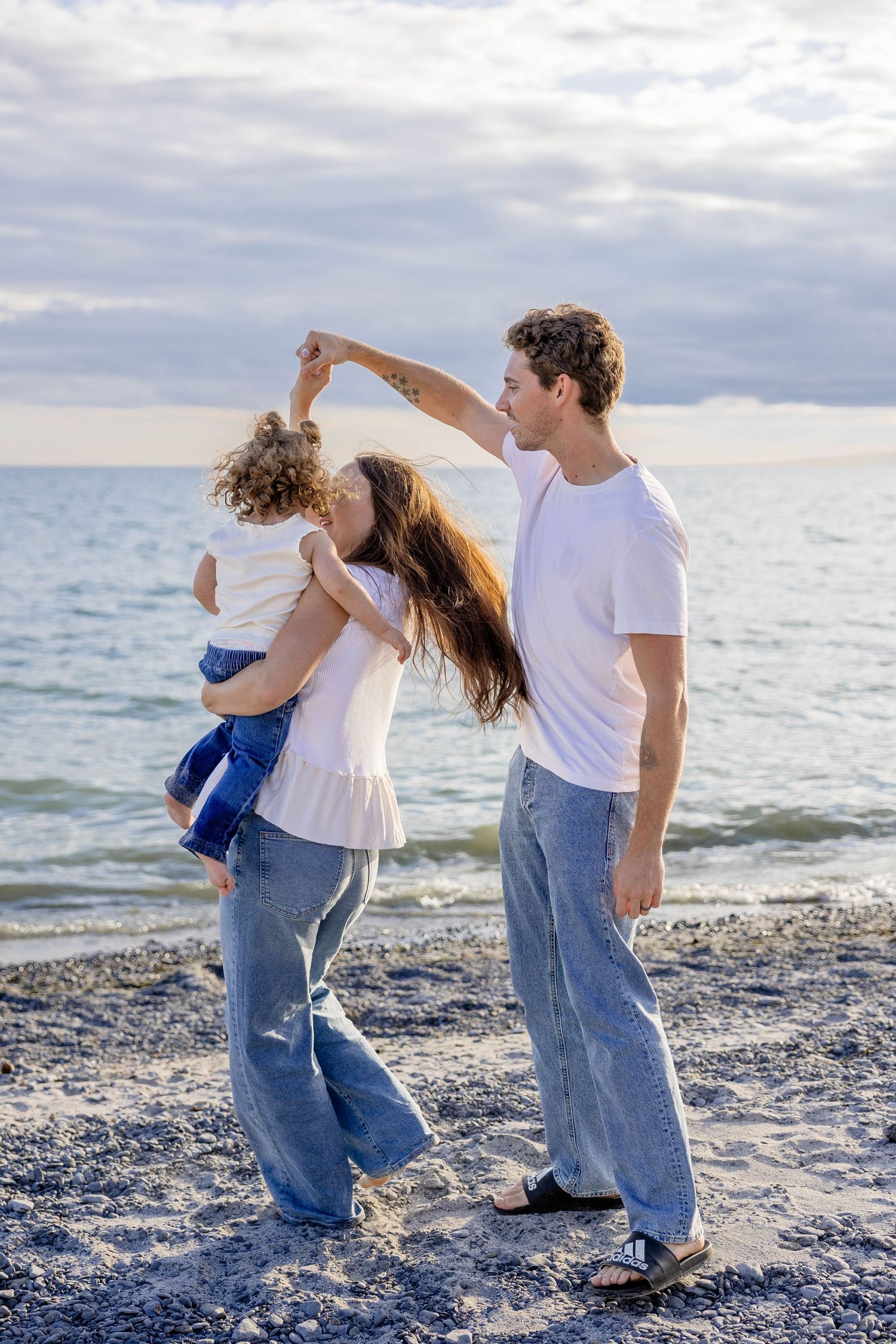 Happy family dancing on a rocky beach, parents holding toddler, wearing white tops and jeans by the ocean.