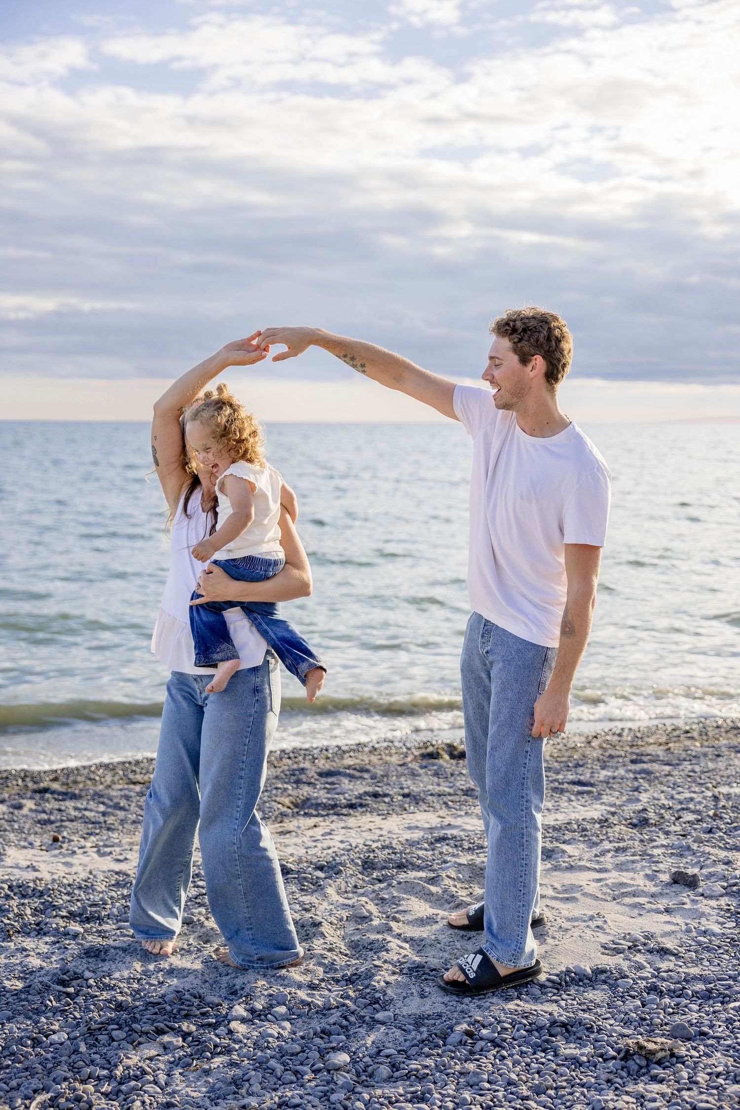 Joyful family dancing on rocky beach, parents spinning with child in arms near ocean shoreline.