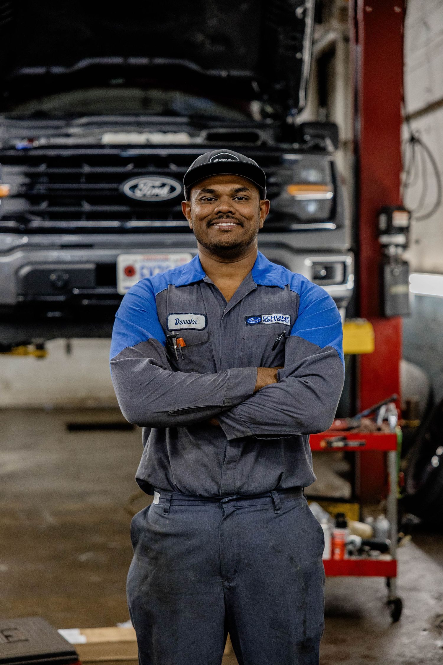 Smiling Ford mechanic in blue uniform stands with arms crossed in front of a Ford truck in an auto repair shop.