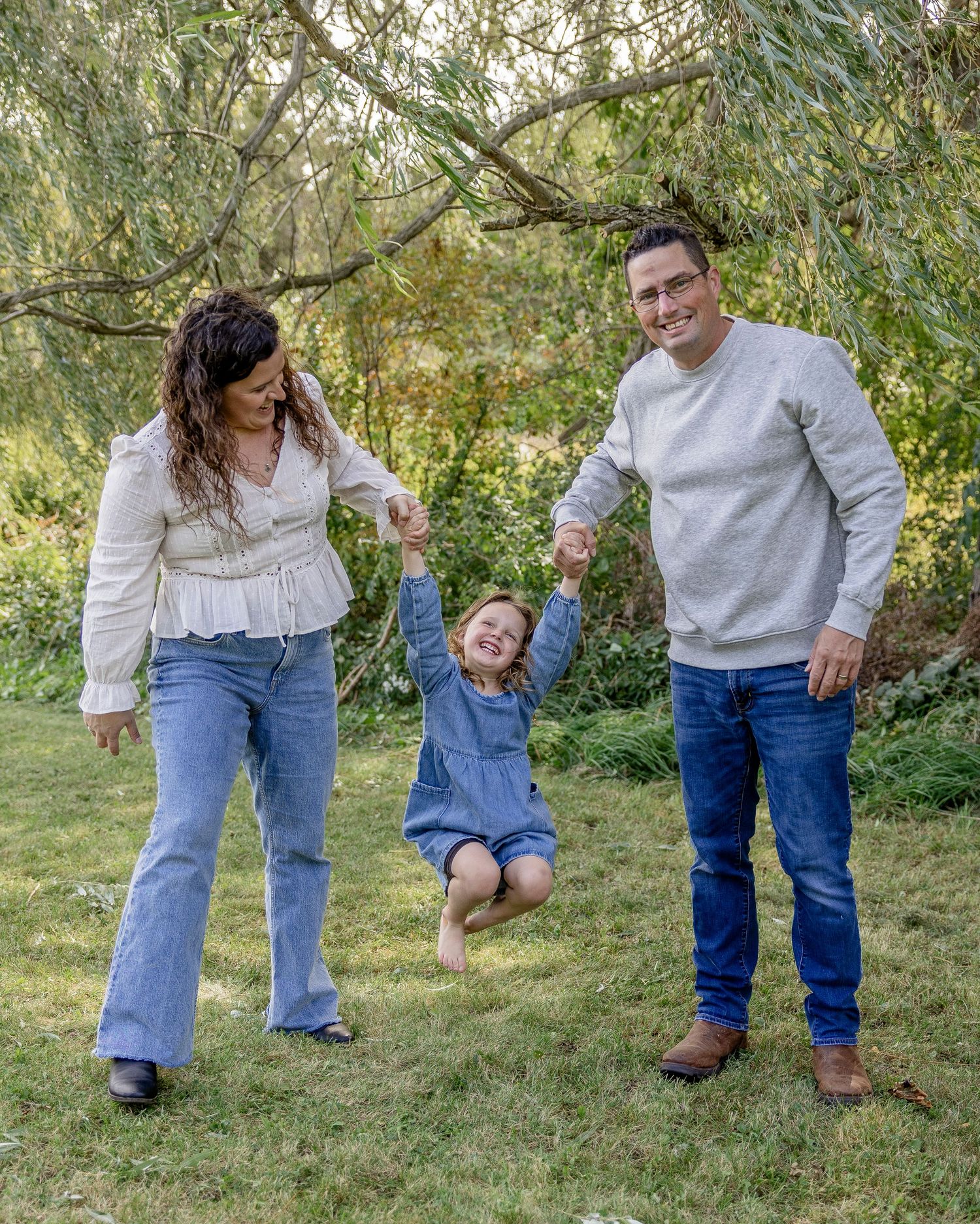 Family of three outdoors, parents swinging smiling child between them under willow tree on green lawn.