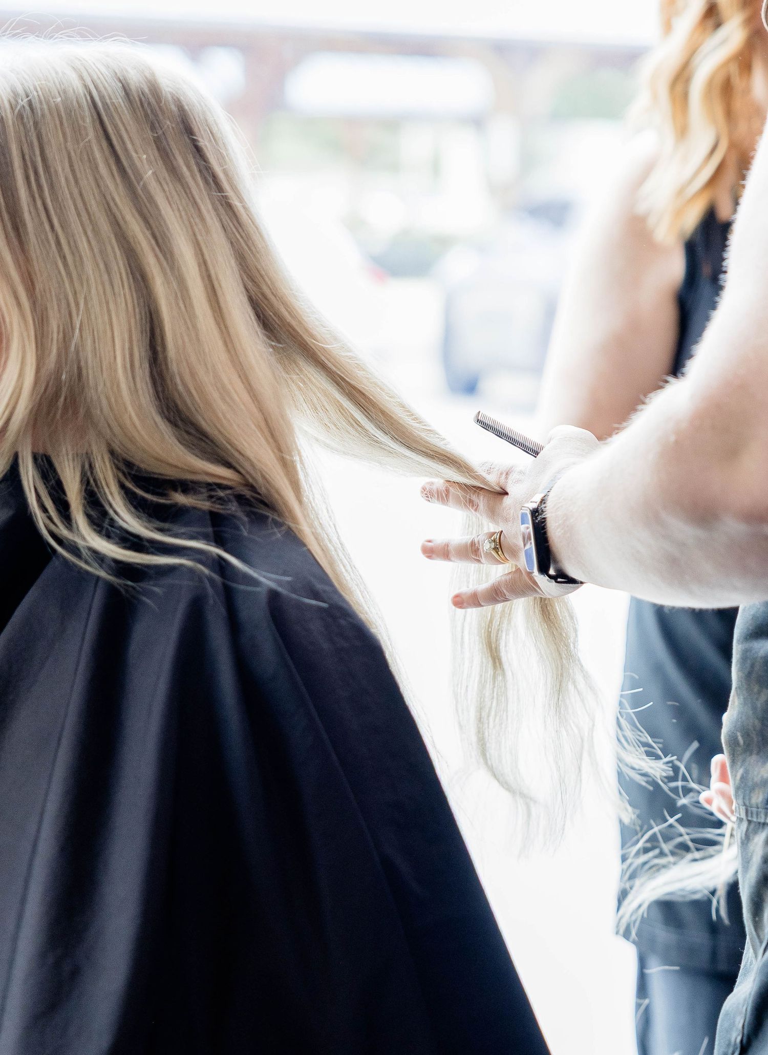 Hair stylist trimming long blonde hair of a client wearing a black salon cape.