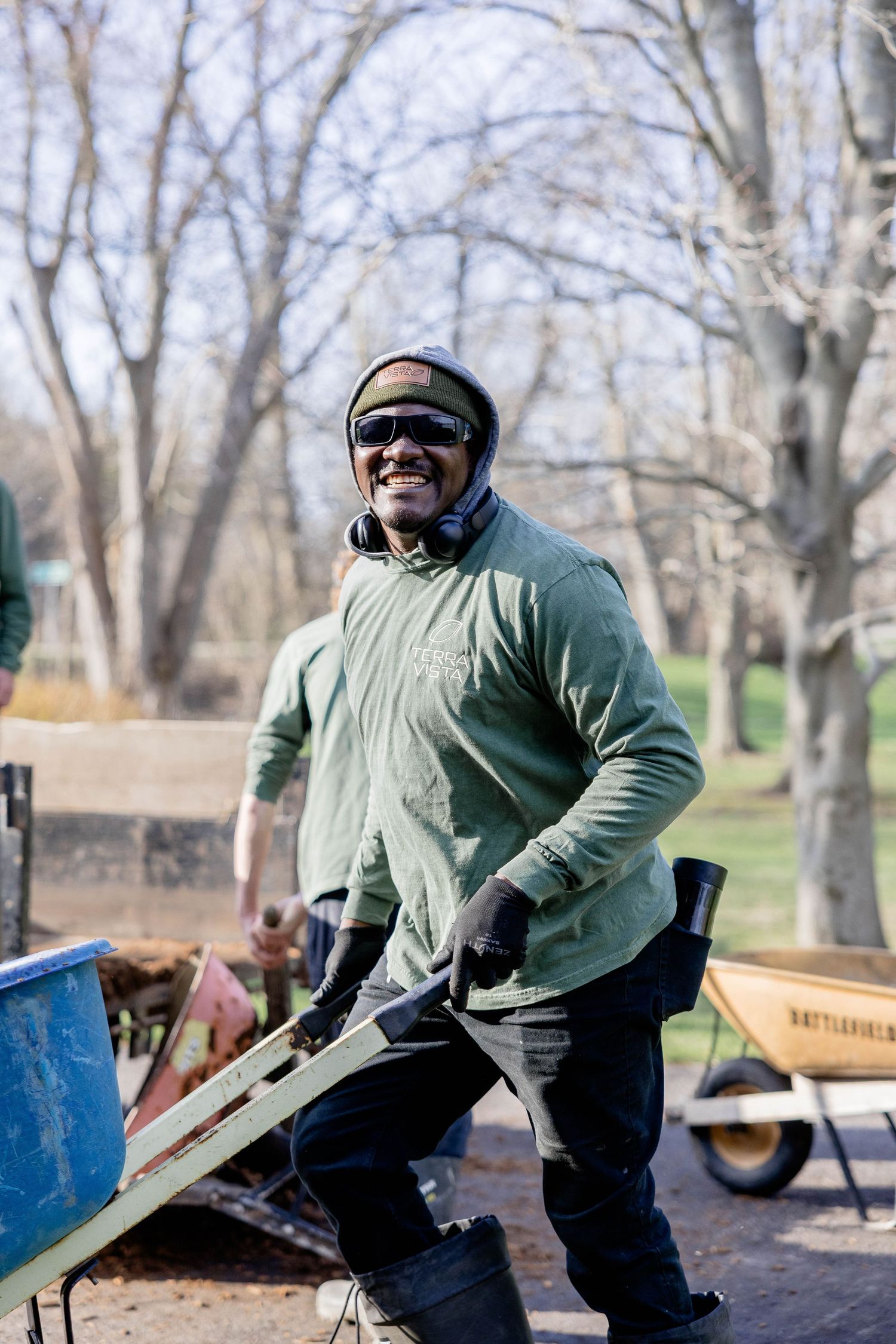 Smiling worker in green shirt and safety goggles pushes a wheelbarrow at an outdoor worksite.