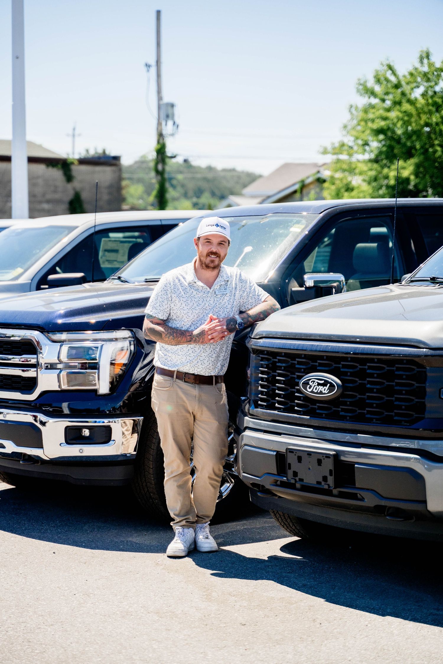 Man in white polo and khakis leaning against Ford F-150 trucks at a dealership lot.