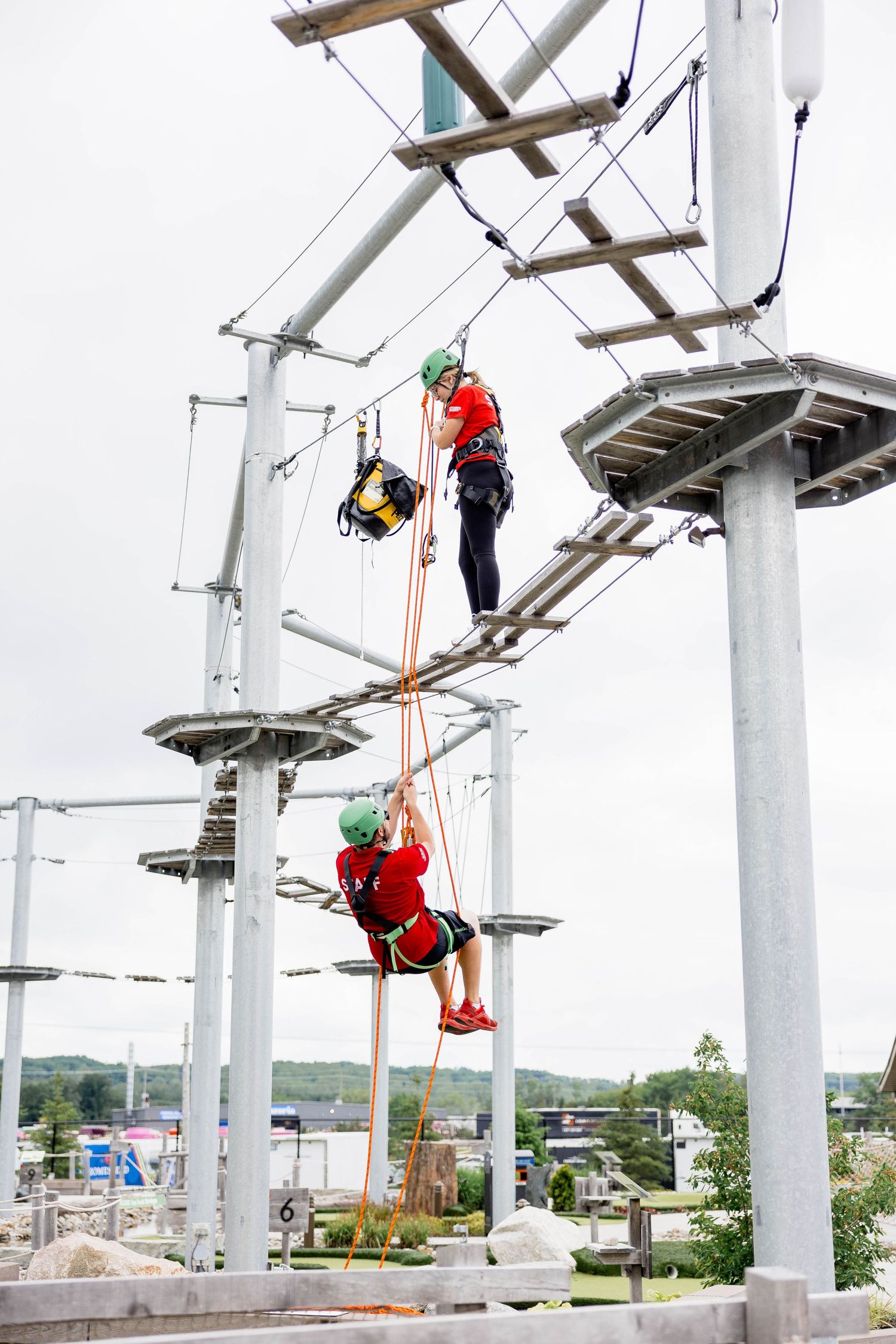 Two climbers in red shirts and green helmets navigate a high ropes adventure course on metal poles.