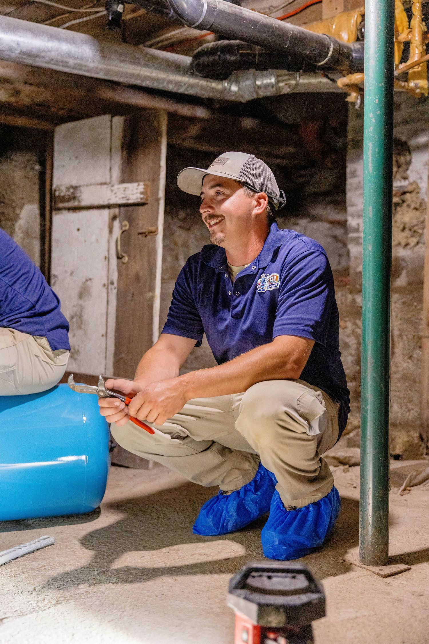 A1 Plumbing technician crouching in basement inspecting blue pressure tank installation.