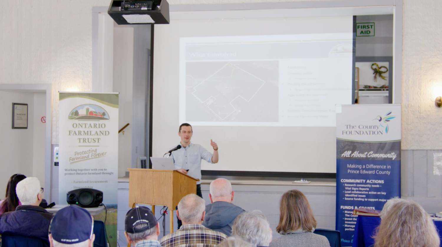 Speaker presenting at a podium to an audience in a bright community hall with projected slides.