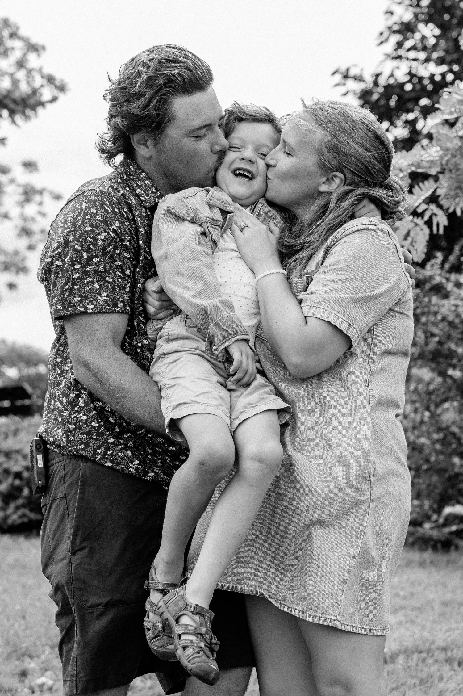 Black and white photo of parents kissing their laughing toddler on the cheeks outdoors.