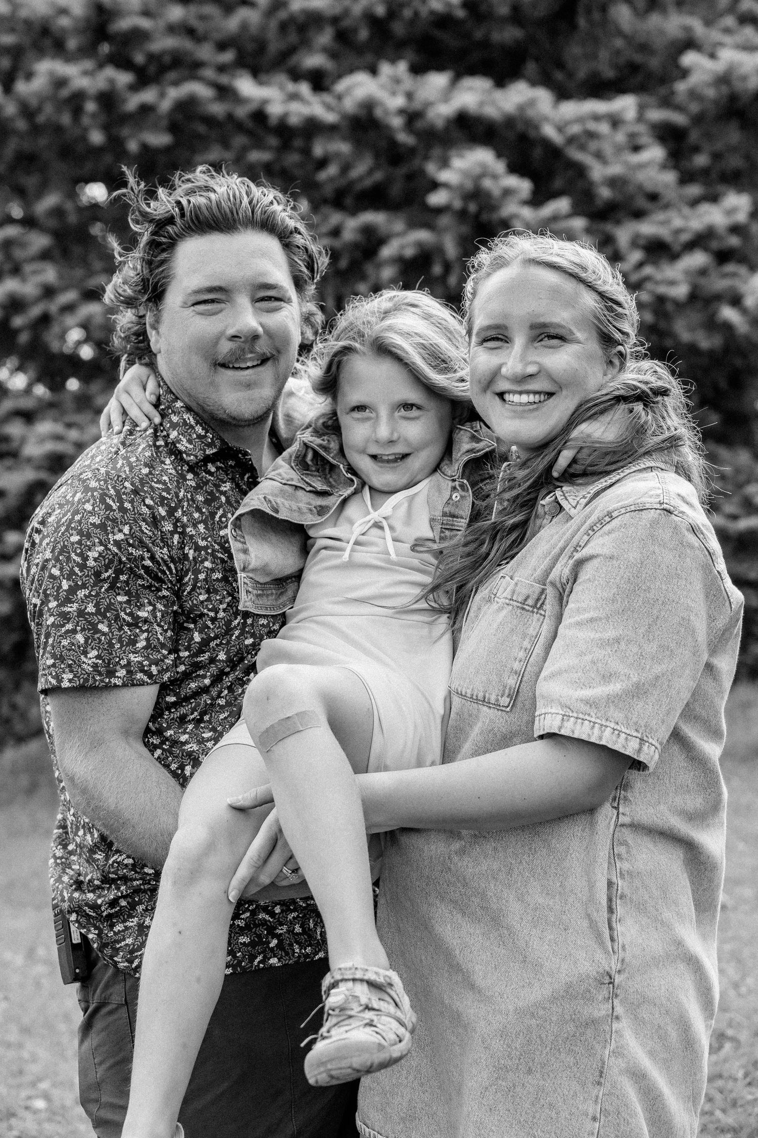 Black and white family portrait of two smiling parents holding their young daughter outdoors near lush green trees.
