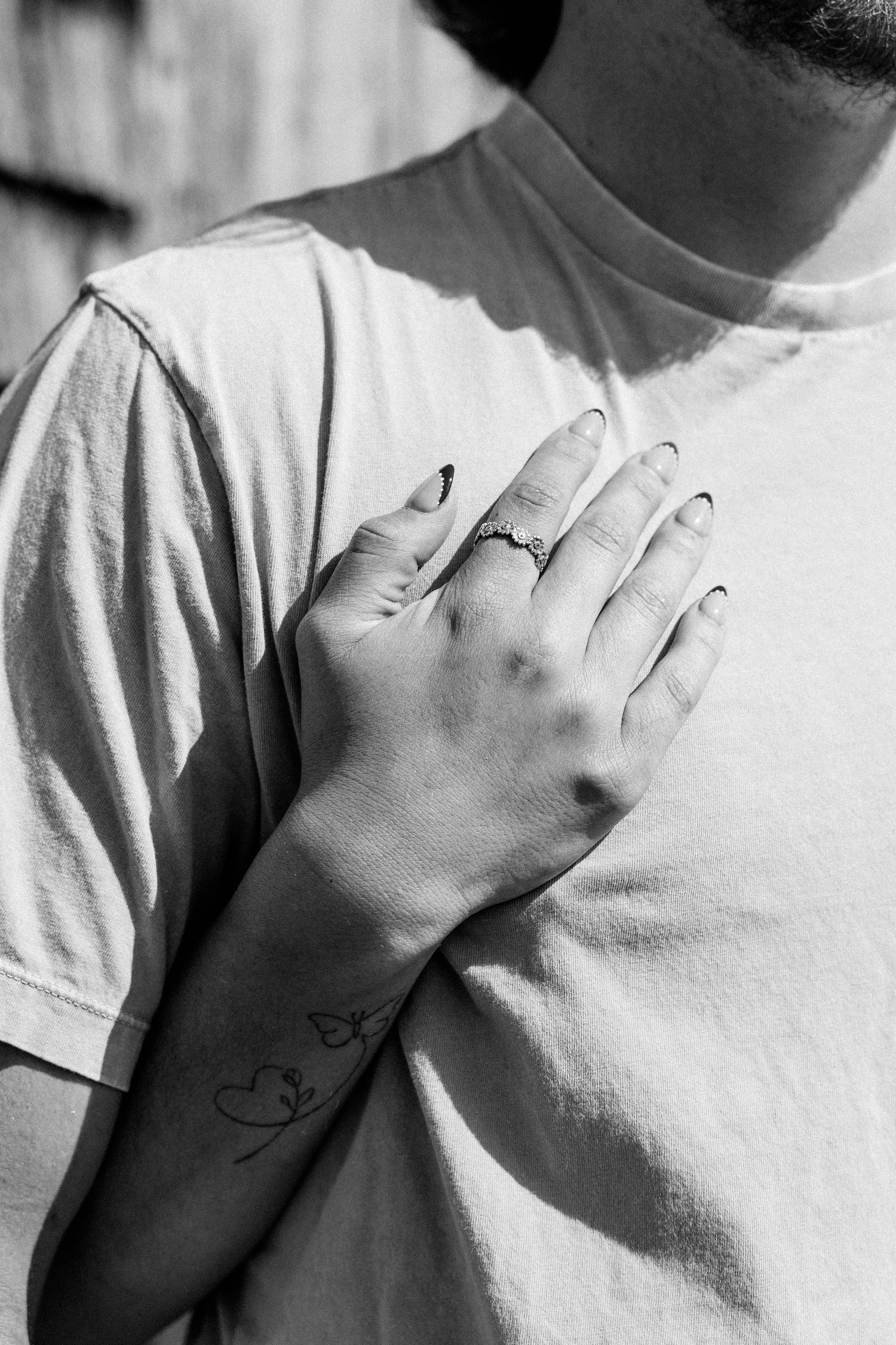 Black and white photo of a hand with black nails, a ring tattoo, and a small heart tattoo on the wrist.