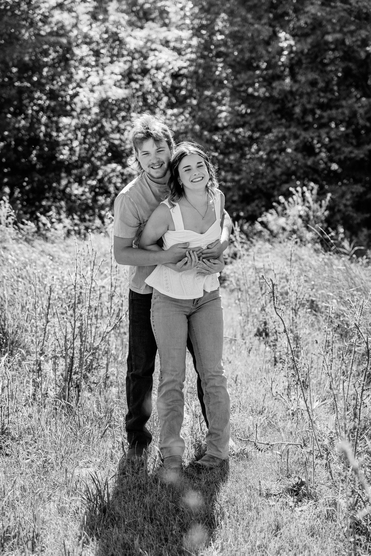 Black and white photo of a happy couple embracing and smiling in a sunny meadow with trees in the background.