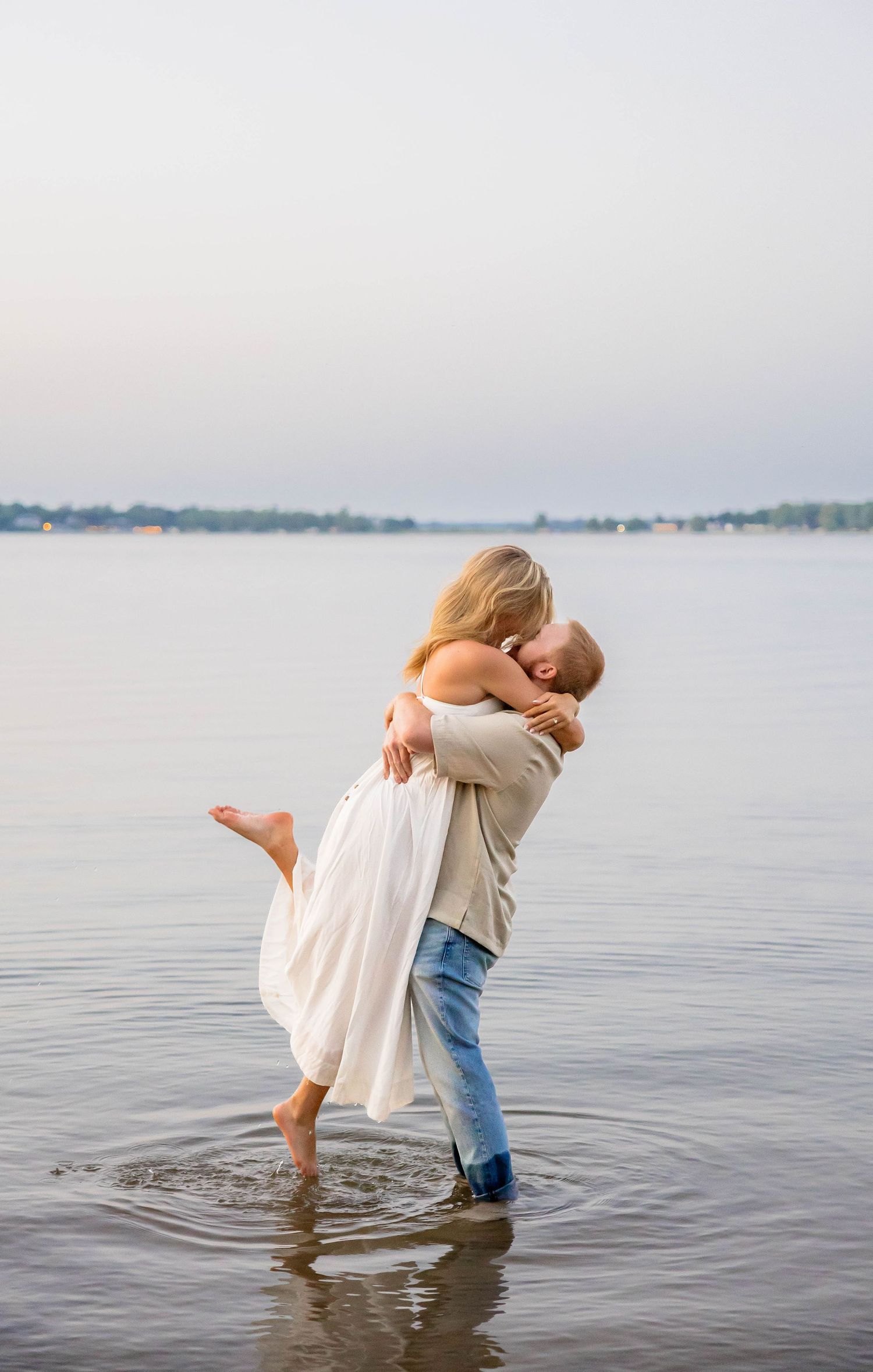 Couple embracing and kissing in shallow water, woman in white dress lifted by man in jeans, romantic lakeside moment.