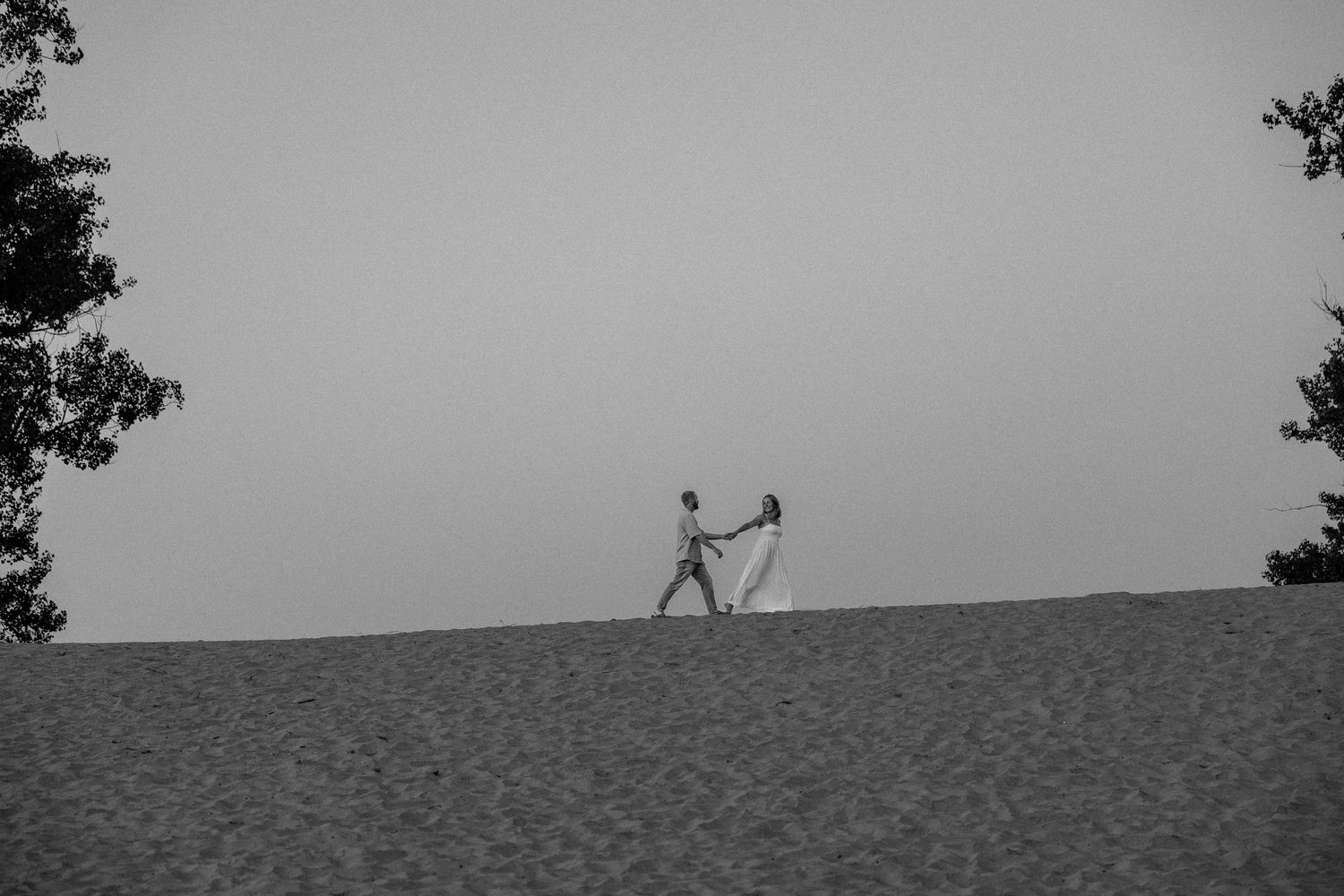 Black and white photo of a couple holding hands on a sandy dune, framed by trees against a misty sky.