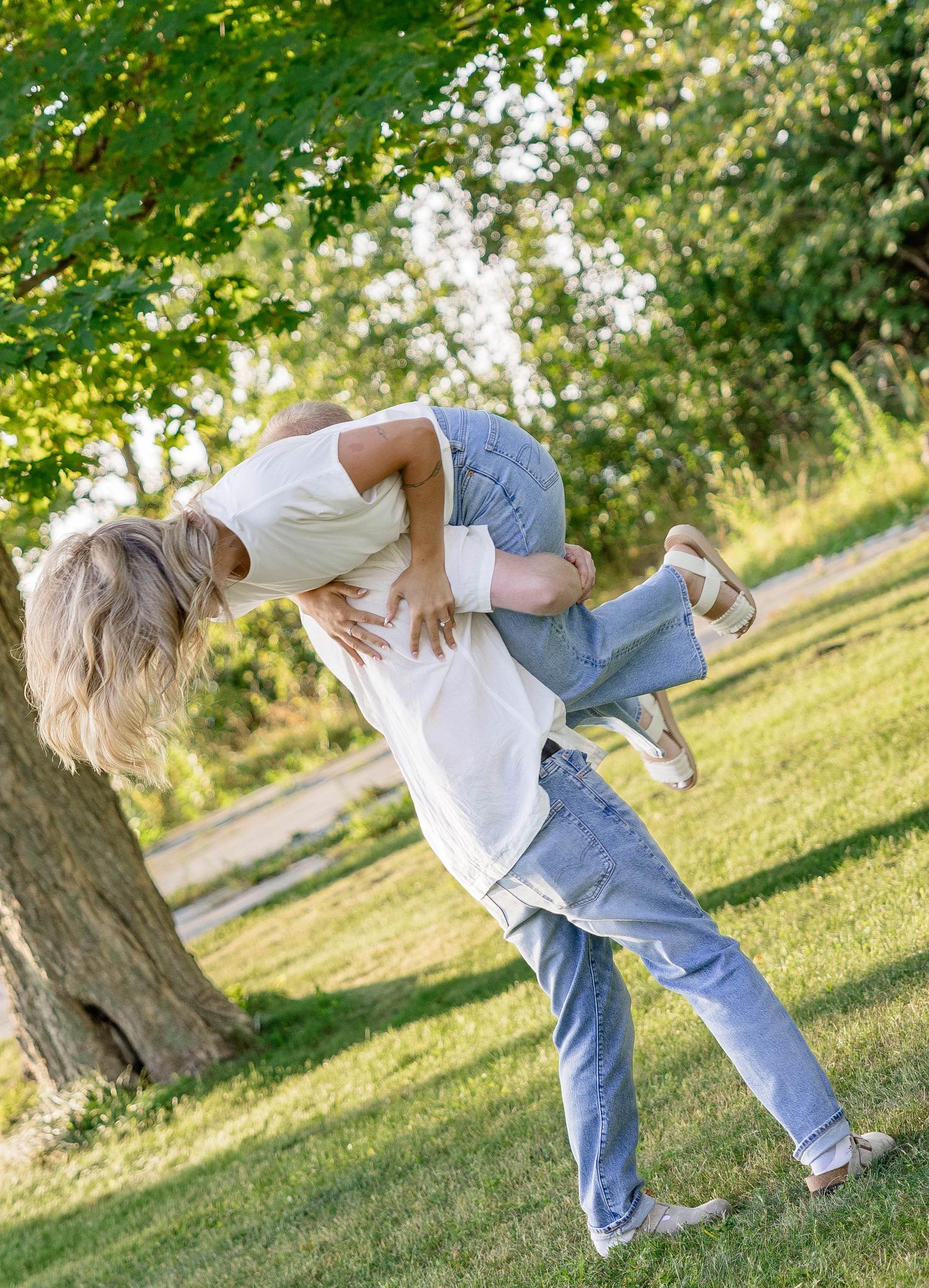 A person playfully lifts another in a sunny green park, both wearing white shirts and jeans, laughing outdoors.