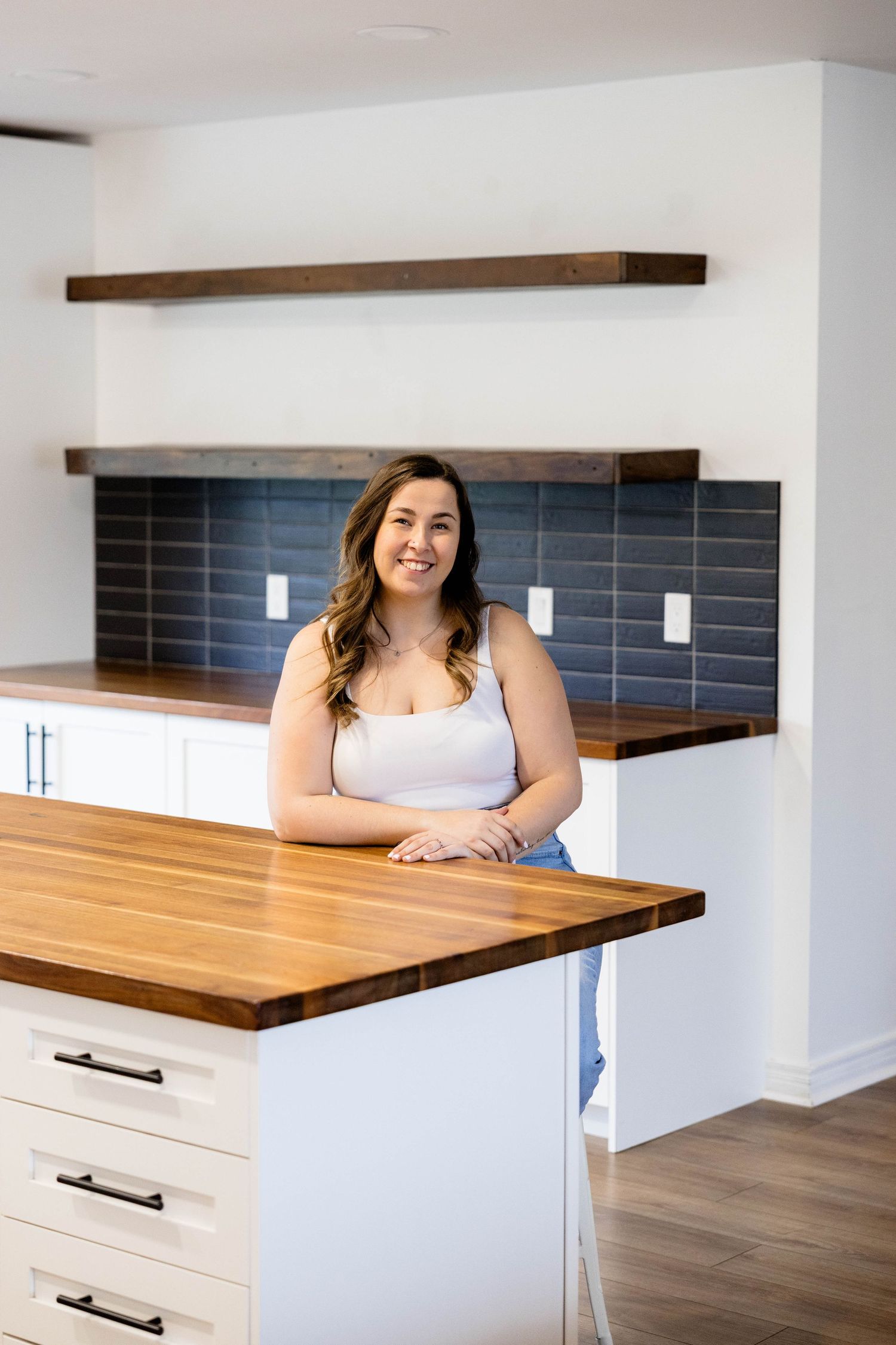 Young woman smiling at kitchen island with butcher block countertop, white cabinets, and blue tile backsplash.