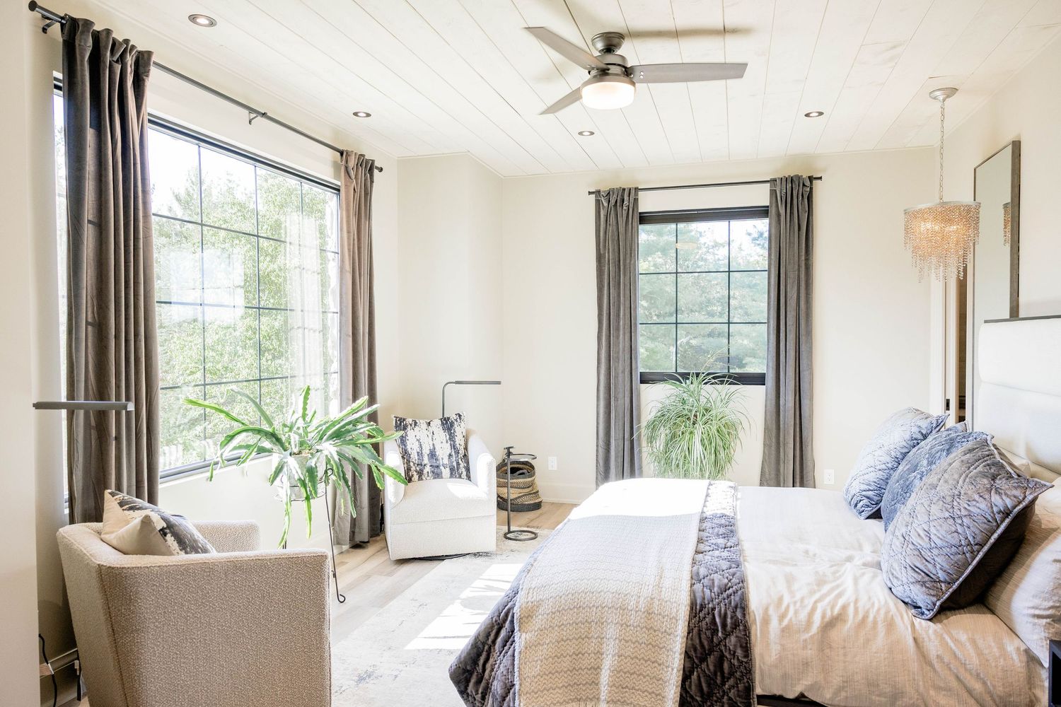 Bright airy bedroom with large windows, blue accent pillows, ceiling fan, and light wood floors.