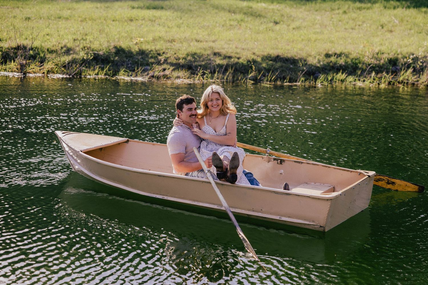 Couple smiling and sitting together in a wooden rowboat on a calm green lake.