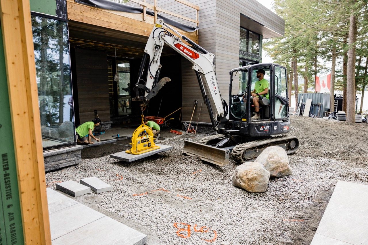 Bobcat mini excavator with yellow MQUIP vacuum attachment lifting stone slabs at a modern home construction site.
