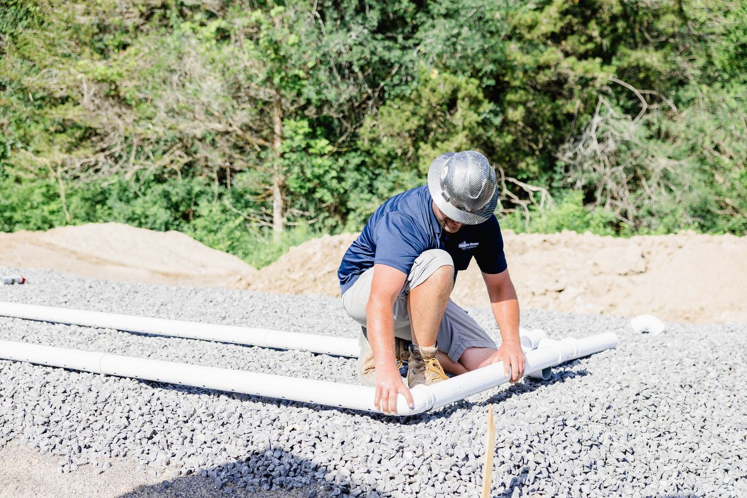 Plumbing technician in hard hat installing underground pipe in gravel foundation trench.
