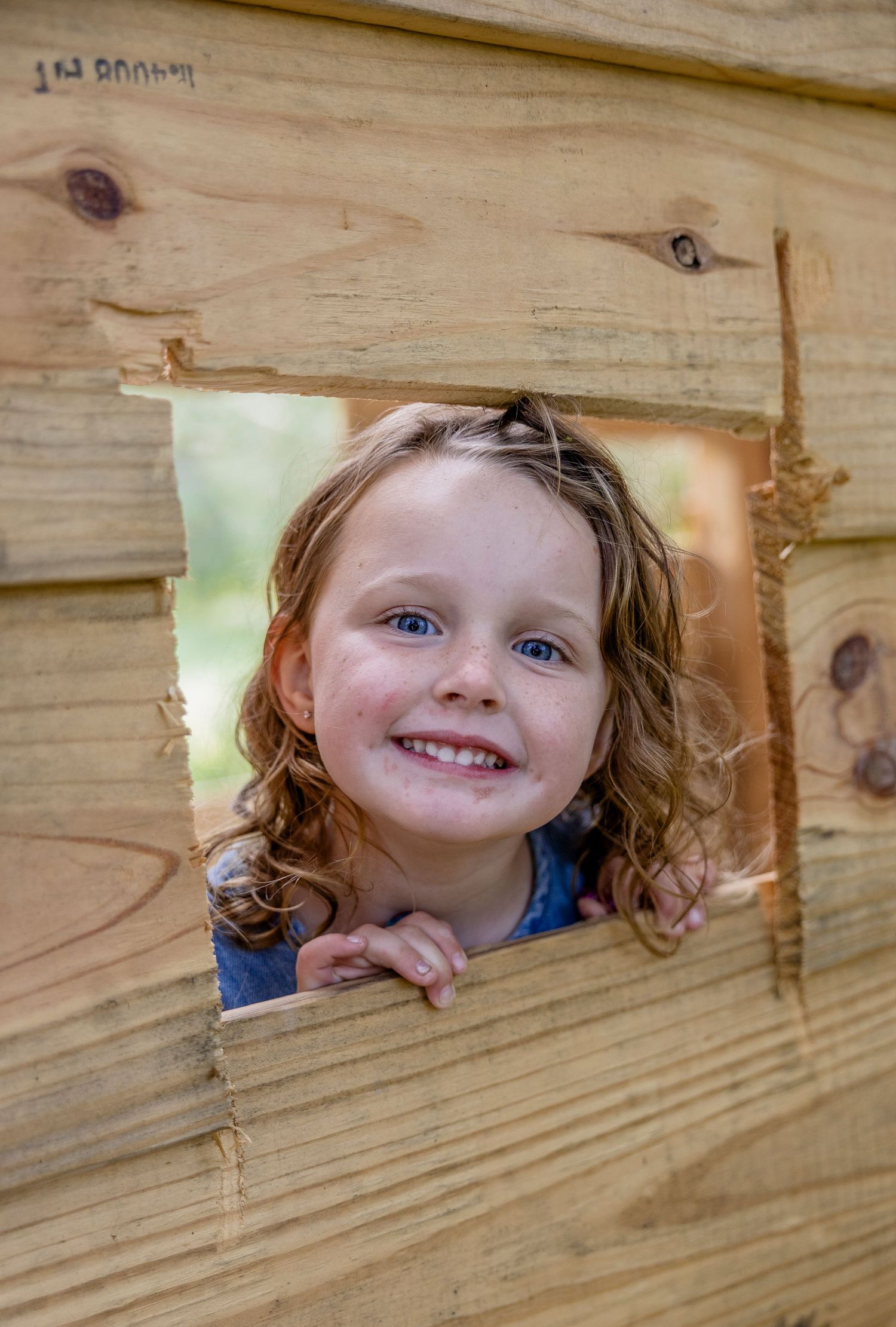 Smiling young girl with curly hair peers through a square wooden window cutout in a rustic playhouse.